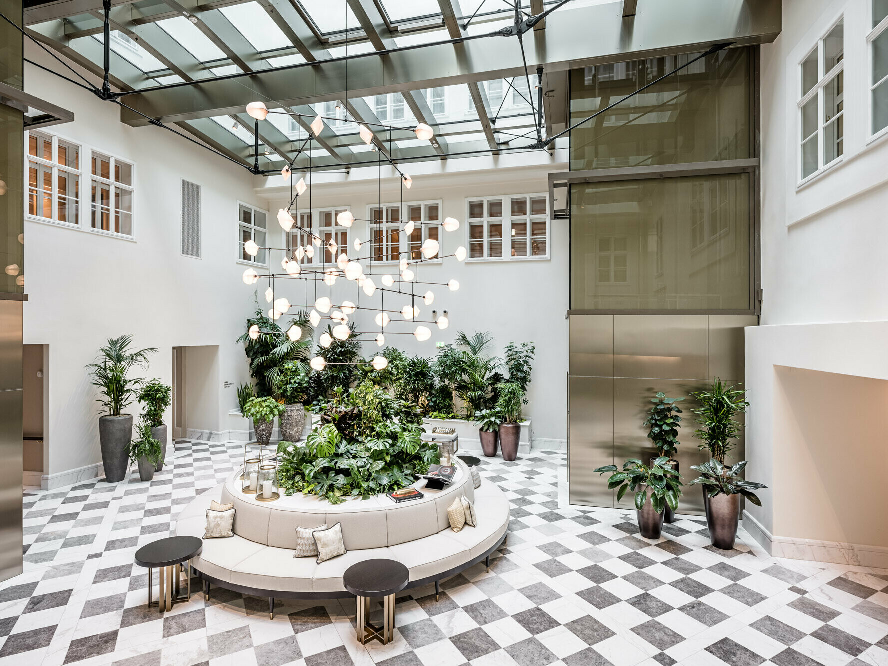 Interieur des Luxushotels, in dem Bronze, Grau und Weiß überwiegen: Auf einem Marmorboden mit Schachbrettmuster befinden sich Sitzgelegenheiten und größere Topfpflanzen, eine moderne Beleuchtung hängt von der Decke in den Raum hinein.