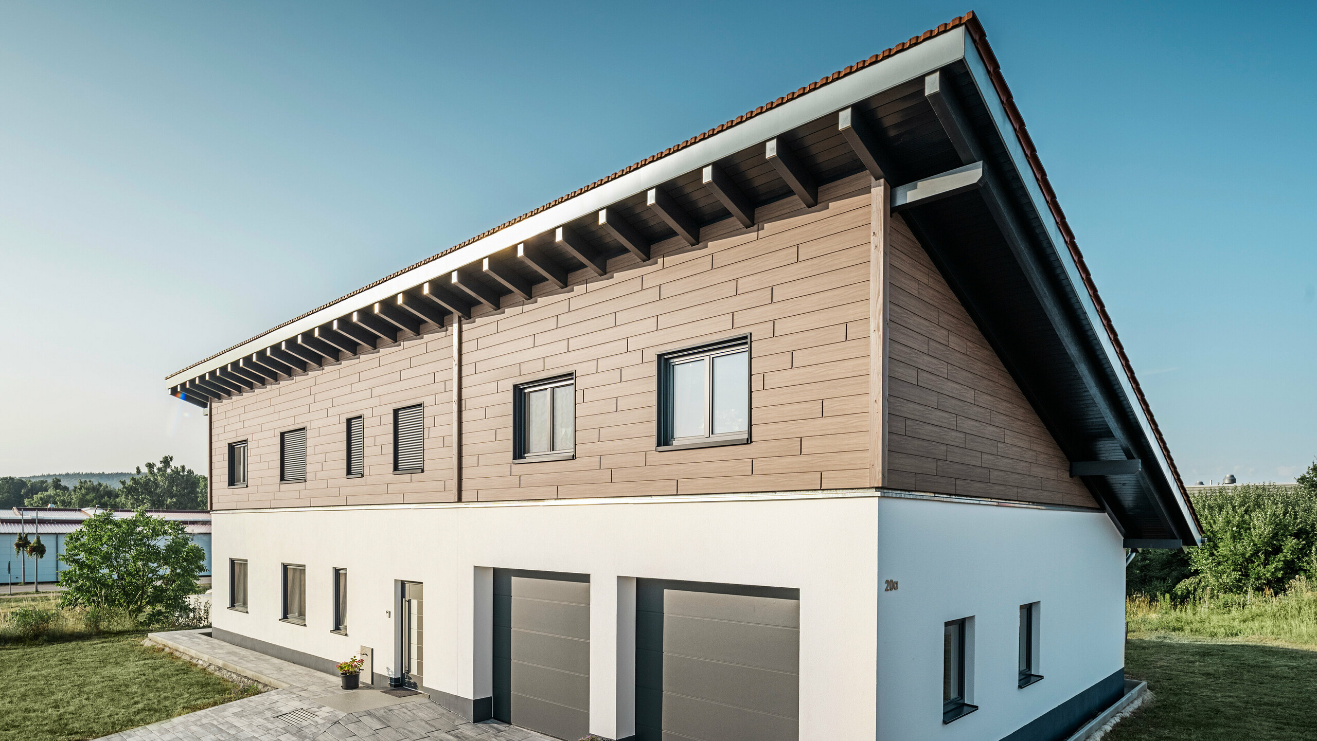 Blick von der Seite auf ein Einfamilienhaus mit Pultdach in Bruck. Das obere Stockwerk ist mit PREFA Sidings in Walnuss Braun ummantelt. Auf der rechten Seite des Bildes sind zwei Garagentore in Anthrazit zu sehen. In der Gebäudemitte befindet sich die Eingangstüre. Der Sockel ist in einem schlichten Weiß gehalten. 