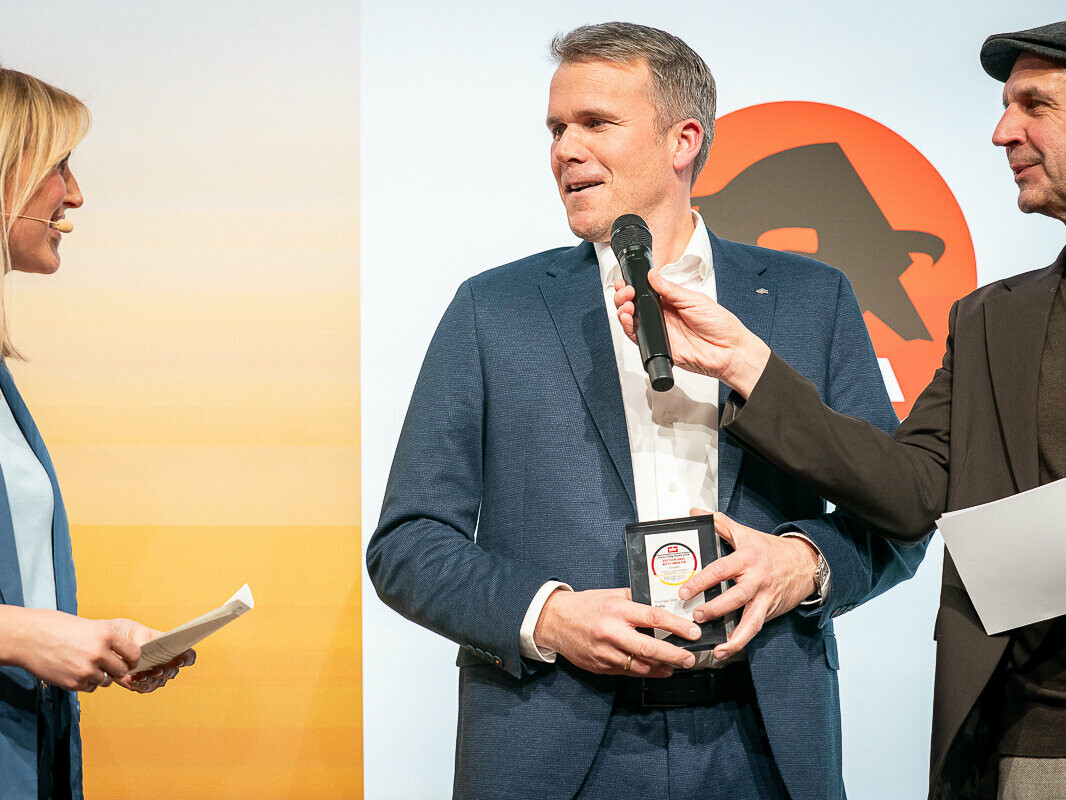 Tobias Götz, Geschäftsführer von PREFA, steht auf einer Bühne und nimmt beim Life & Living Award 2026 eine Auszeichnung entgegen, während er in einem Interview von zwei Moderatoren befragt wird.