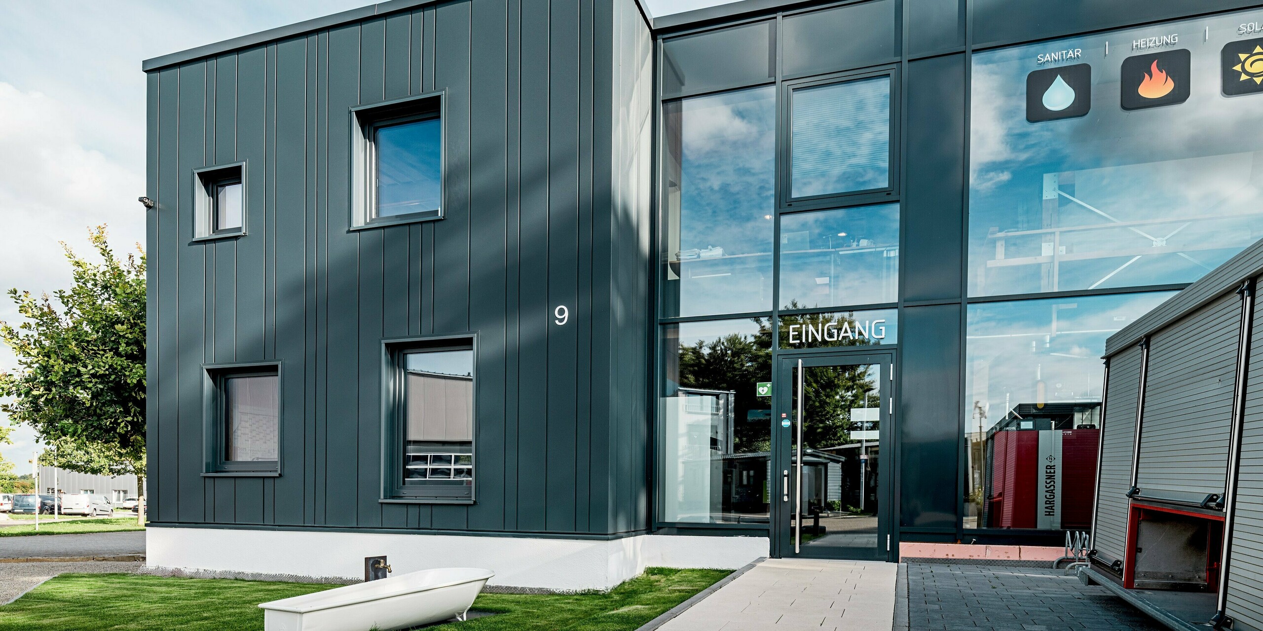 Dieses Bild zeigt den modernen Eingangsbereich der Gebäudetechnik Schädel GmbH mit einer markanten PREFA Aluminiumfassade in P.10 Anthrazit. Die vertikal verlegten Sidings in unterschiedlichen Baubreiten erzeugen eine lebendige Struktur. Durch die reflektierenden Glasflächen entsteht ein dynamisches Zusammenspiel aus Licht und Material. Die robuste PREFA Fassade bietet dauerhaften Schutz und moderne Eleganz.