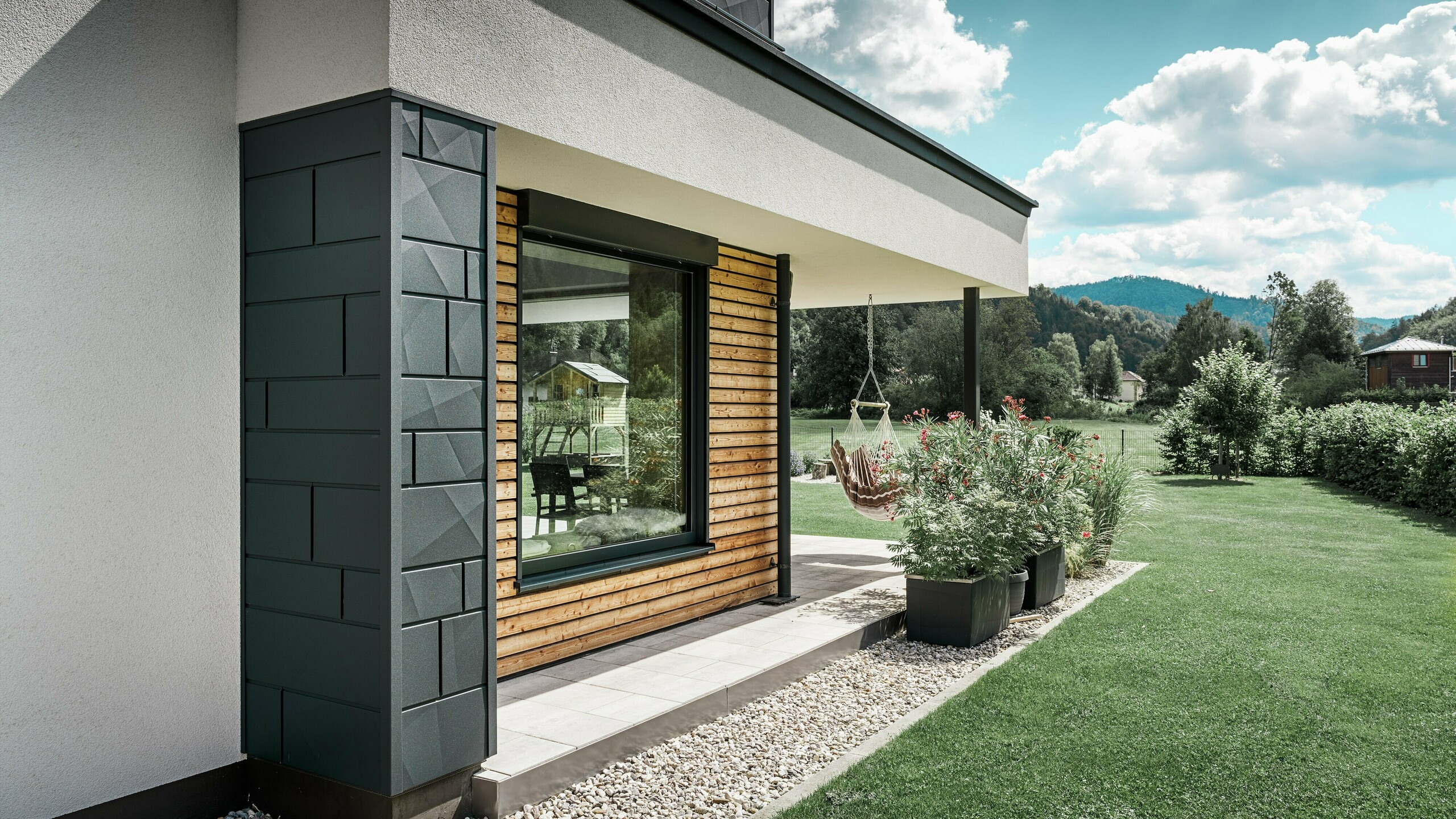 Teilaufnahme eines neu gebauten Einfamilienhauses. Hier wurden an der Fassade unterschiedliche Materialien verwendet. Zur weißen Putzfassade werden Highlights mit den PREFA Aluminium Siding.X in Anthrazit sowie horizontalen Holzpaneelen, welche ein großes Fenster umfassen, gesetzt. Auf der Terrasse hängt ein Hängesessel.