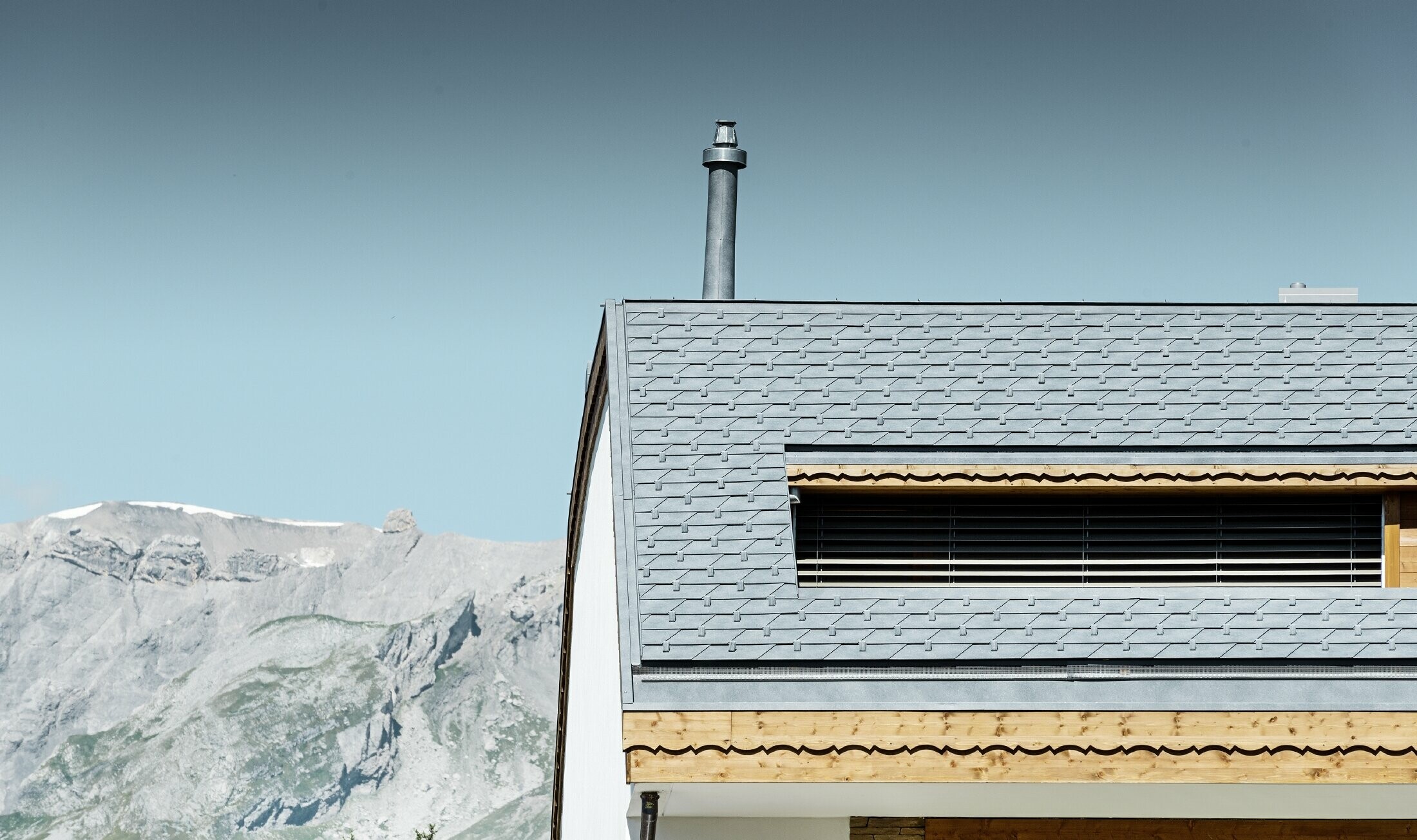 Mehrfamilienhaus in Crans-Montana mit Bergen im Hintergrund und einer Fassade mit verspielten Holzelementen mit einem PREFA Aluminium Dachschindel-Dach in steingrau 