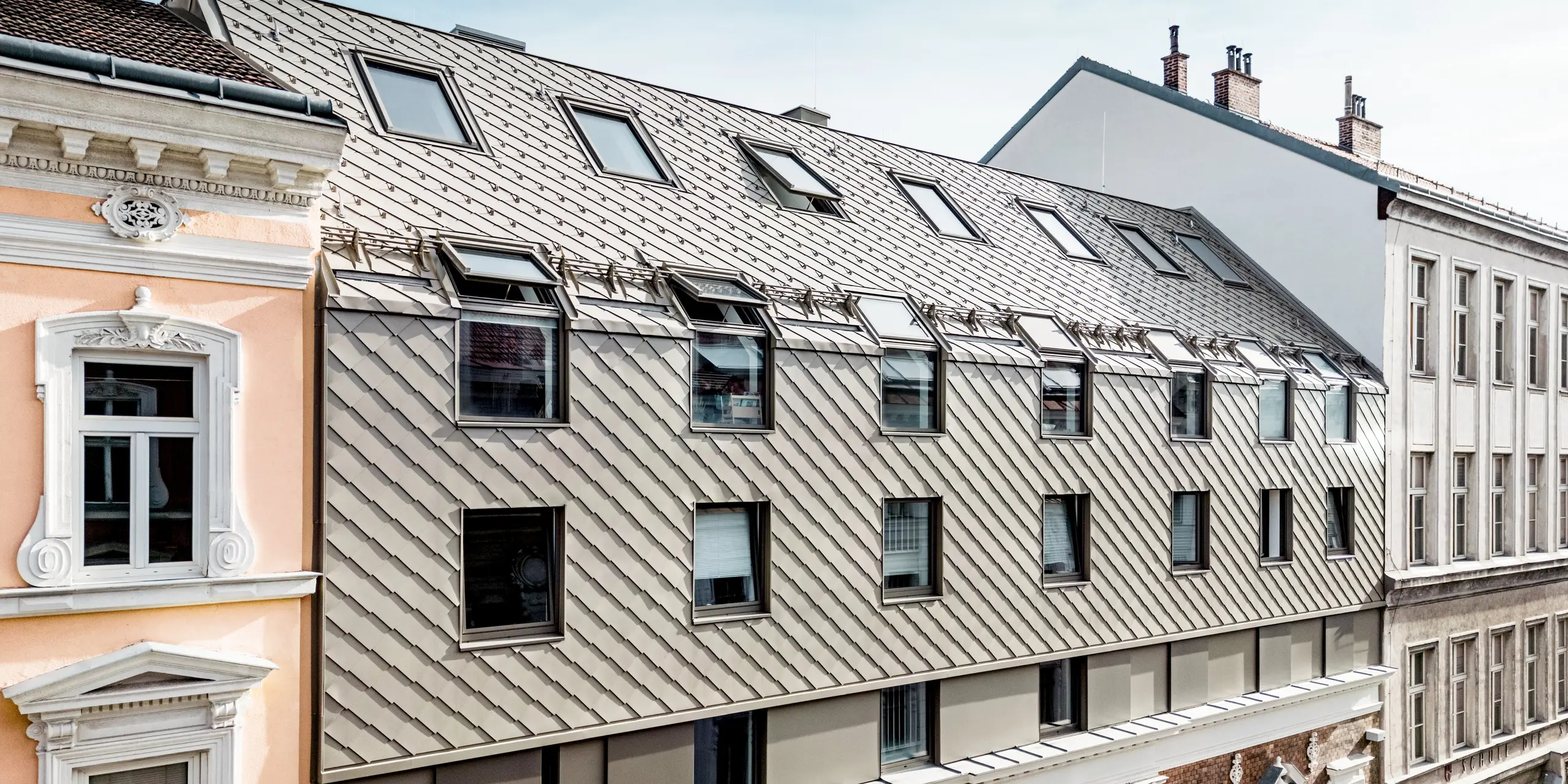 Das Bild zeigt die prägnante Erweiterung eines Wiener Wohngebäudes, bei dem moderne PREFA Rauten 29×29 in Bronze die obere Etage zieren. Die Rauten verleihen der Architektur eine elegante und zeitgemäße Note, die perfekt mit der historischen Fassade darunter harmoniert. Besonders auffällig sind die Dachgauben, die das Dach beleben und eine interessante Struktur schaffen. Der Kontrast zwischen der klassischen Ziegelmauer und den modernen Aluminium-Rauten betont die gelungene Mischung aus Tradition und Innovation in der Wiener Architektur.