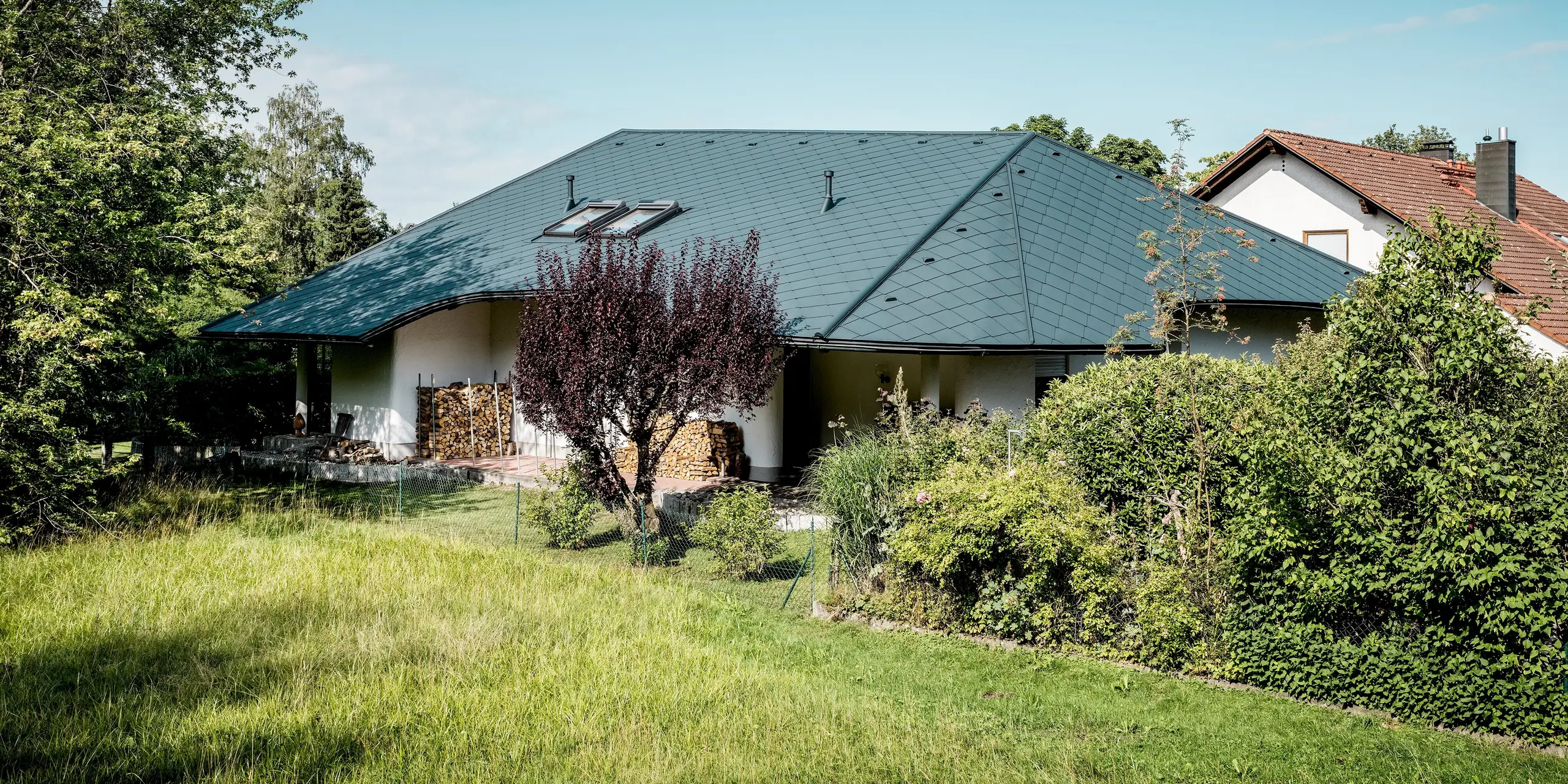 Weitläufige Gartenansicht des sanierten Einfamilienhauses in Bobingen: Die großzügigen, unterschiedlich geneigten Dachflächen mit PREFA Dachraute 44 × 44 in P.10 Anthrazit prägen das Gebäude eindrucksvoll. Dachfenster, geschwungene Trauflinien und die klare Rautenstruktur setzen markante Akzente. Das leichte, robuste Aluminium unterstützt langlebige, witterungsbeständige Lösungen auch bei anspruchsvollen Dachformen.