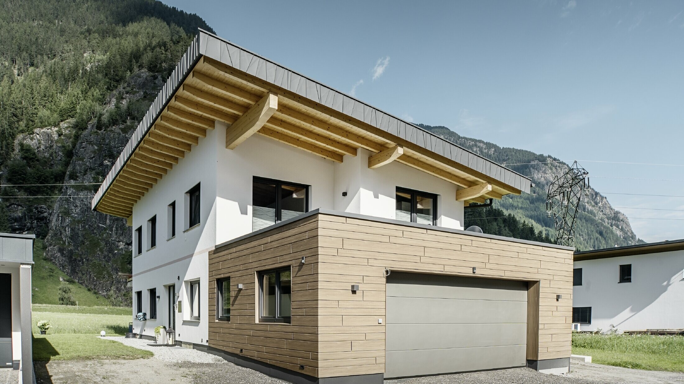 Modernes Einfamilienhaus mit Fassadensanierung: erneuerte Außenfassade mit horizontaler Fassadenverkleidung in Holzoptik, kombiniert mit weiß verputzten Flächen, integrierter Garagenverkleidung und zeitgemäßer Architektur in alpiner Umgebung.