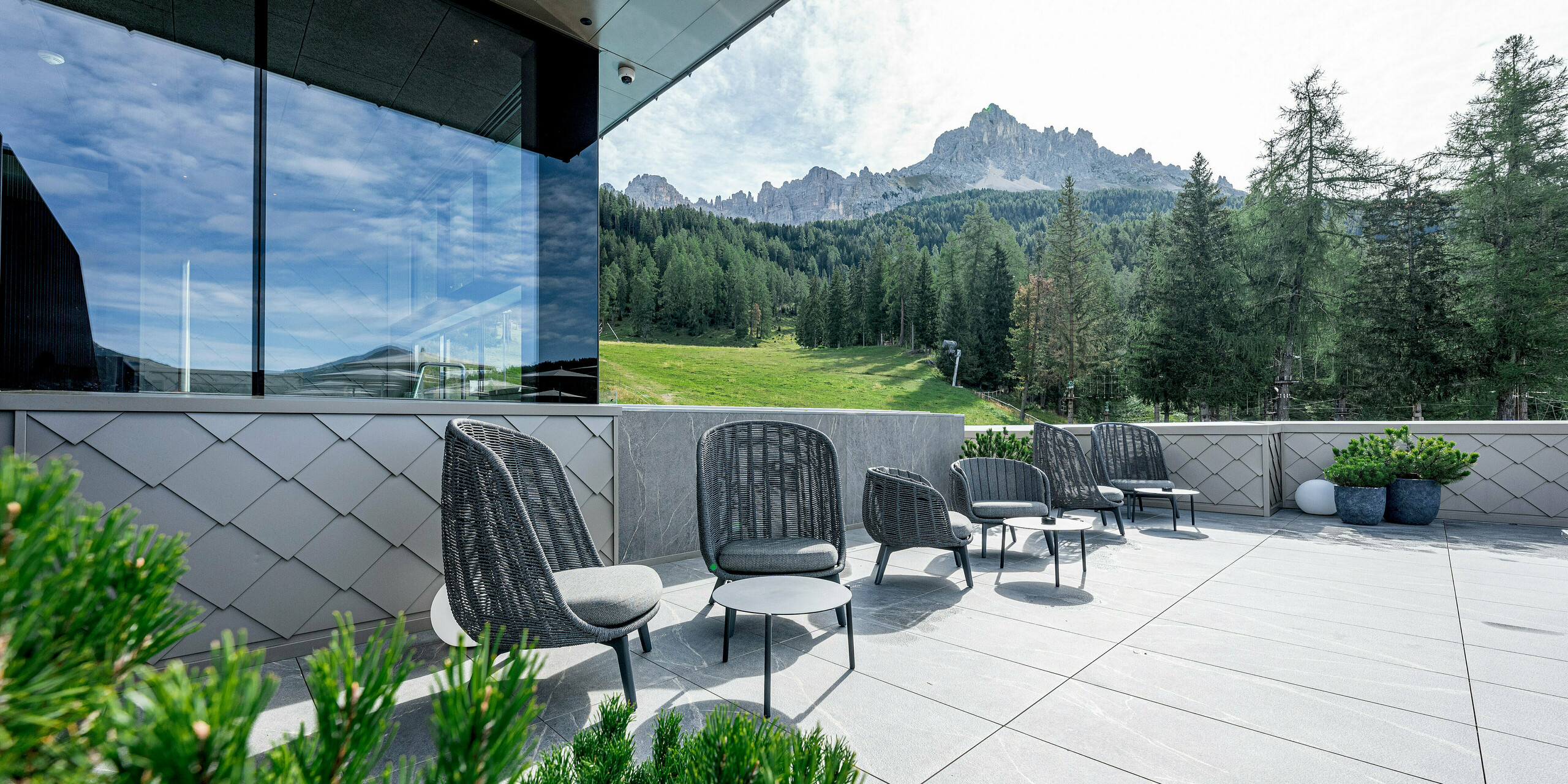 Die Terrasse des Sporthotels Obereggen in Südtirol ist mit modernen Sitzmöbeln und großformatigen Steinplatten ausgestattet und bietet einen weiten Blick auf das alpine Bergpanorama. Die PREFA Aluminiumfassade mit Wandraute 29 × 29 in Bronze setzt einen eleganten architektonischen Akzent. Die fein gezeichnete Rautenstruktur wirkt je nach Lichteinfall lebendig und hochwertig. Dank ihrer Langlebigkeit, Witterungsbeständigkeit und Wartungsfreiheit sind PREFA Aluminiumprodukte ideal für exponierte Hotelterrassen im alpinen Raum.