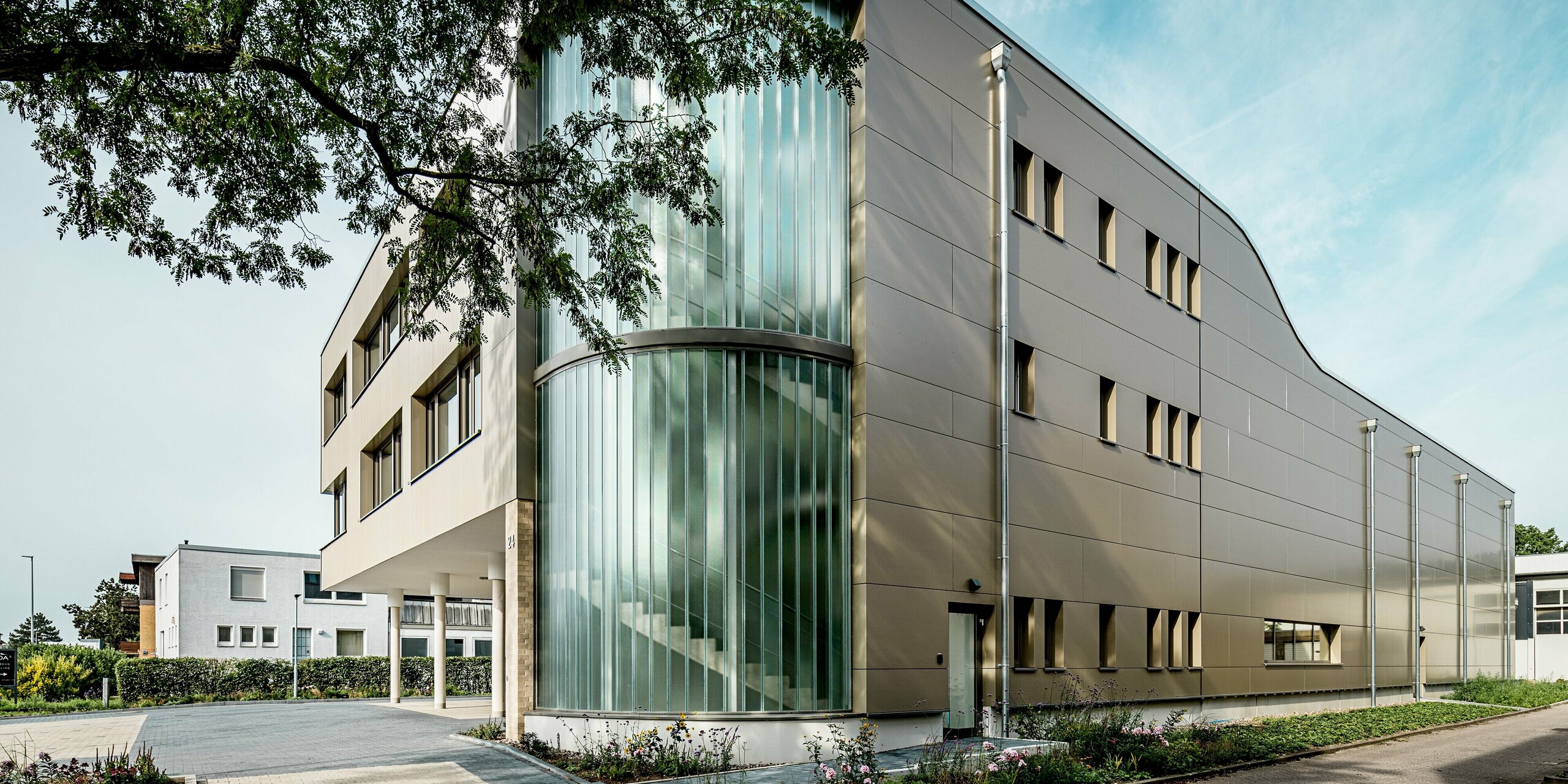 Das Bild zeigt das Festo-Gebäude in Langenhagen mit einer markanten Fassade aus PREFA Aluminium Verbundplatten in einem eleganten Bronzeton. Besonders hervorstechend ist die abgerundete Gebäudeecke, die mit vertikal verlaufenden Glasflächen versehen ist, die das natürliche Licht in das Gebäudeinnere strömen lassen. Diese Glasschneidwand bildet einen auffälligen Kontrast zu den gleichmäßig verteilten Fenstern und der klaren Fassadengestaltung. Der Einsatz von PREFA Aluminiumprodukten sorgt nicht nur für eine ästhetisch ansprechende, sondern auch für eine langlebige und witterungsbeständige Lösung. Die harmonische Verbindung von modernem Design und Funktionalität macht das Gebäude zu einem herausragenden Beispiel für zeitgemäße Architektur.