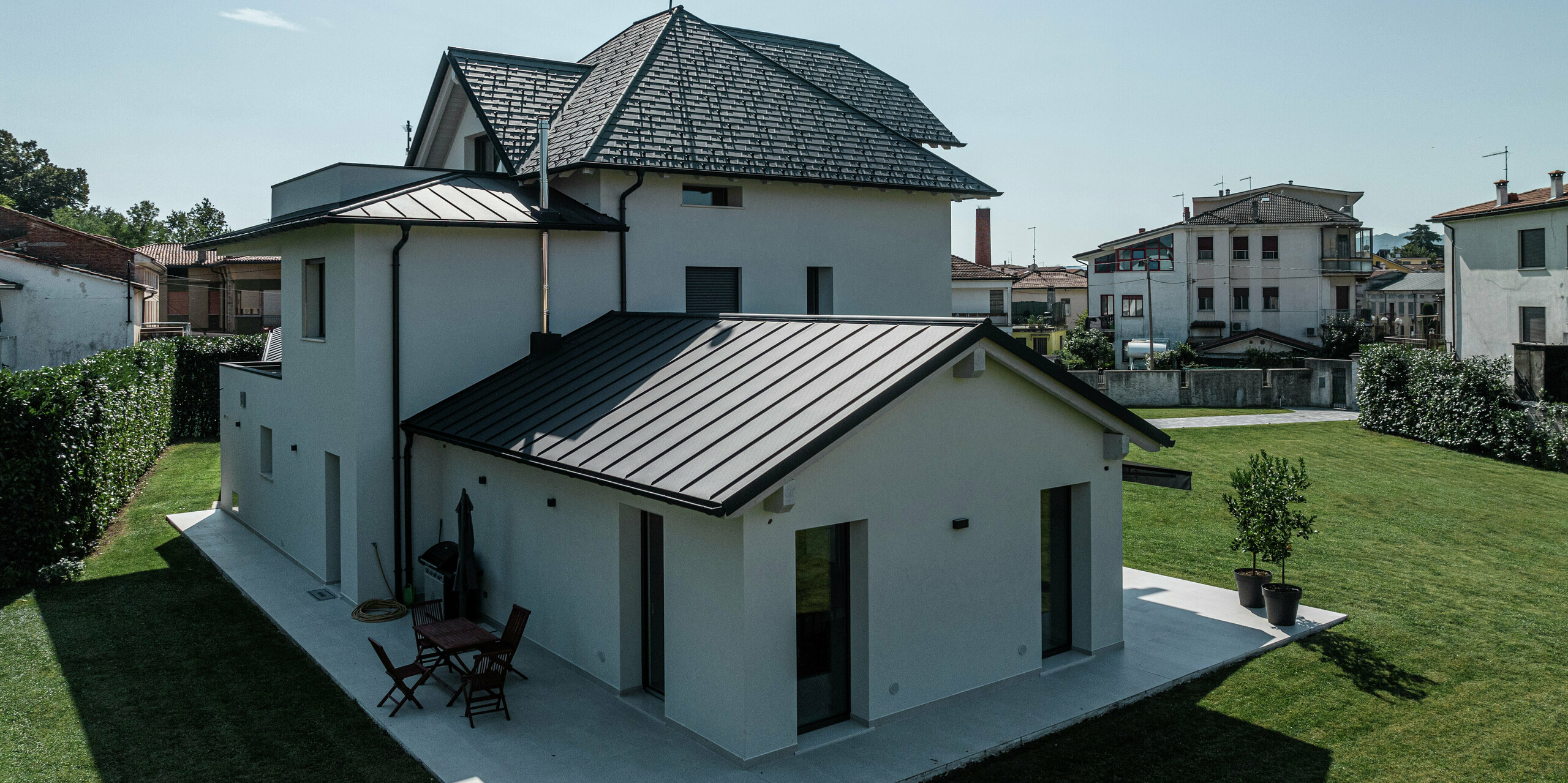 Rückansicht eines zeitgemäßen Einfamilienhauses in Marano Vicentino, Italien, mit einem langlebigen PREFA Aluminiumdach in der hochwertigen P.10-Beschichtung in Anthrazit. Die angrenzenden Nebengebäude sind mit einer robusten Doppelstehfalzdeckung aus Aluminium versehen. Die hochwertige Aluminiumverkleidung verbindet Ästhetik mit Witterungsbeständigkeit.
