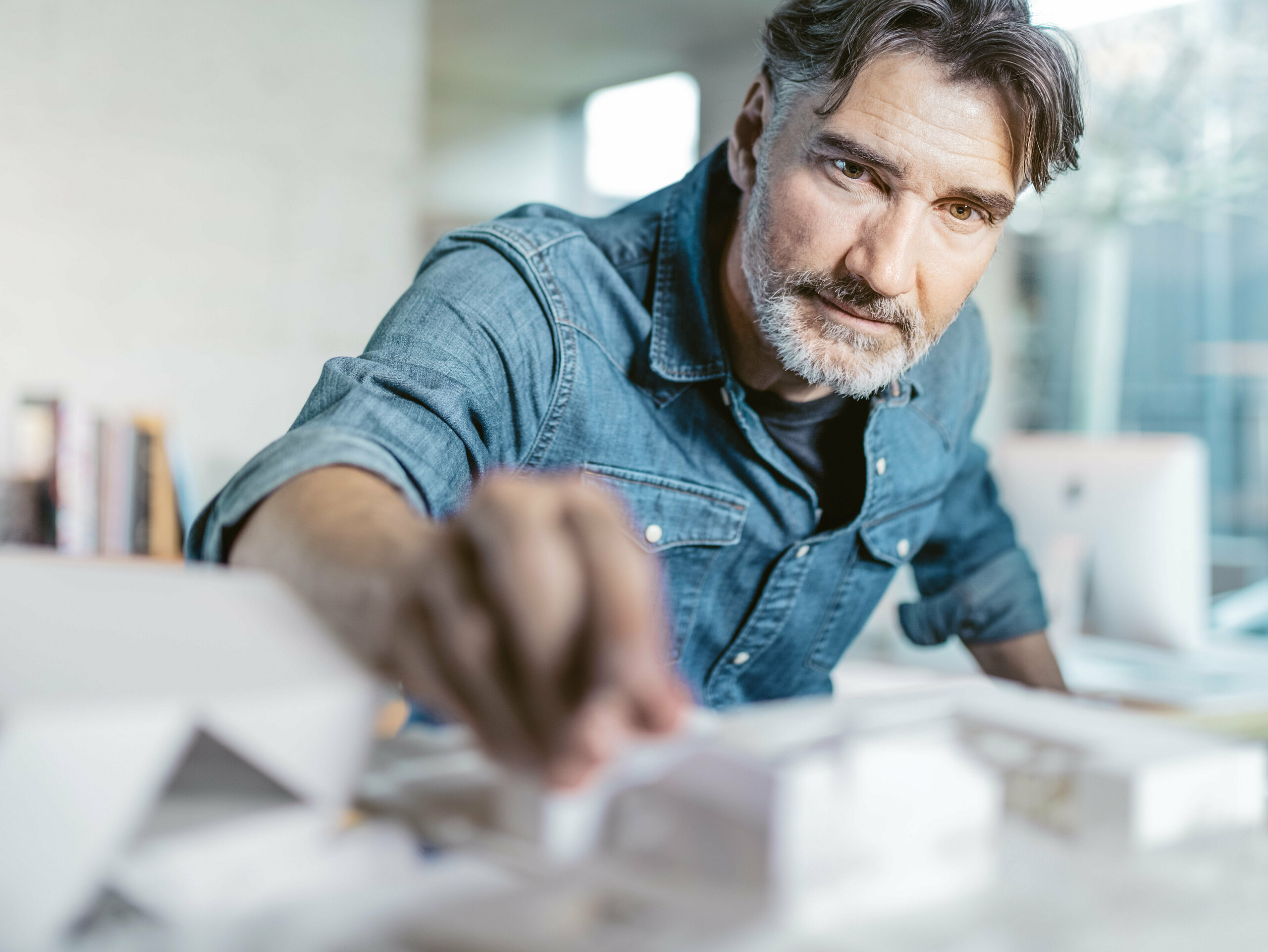 Architekt bei der Arbeit mit einem Architekturmodell, umgeben von Skizzen und Entwürfen, während er die Details eines Projekts prüft. Das Bild vermittelt Kreativität und Präzision, passend zur Bewerbung der hochwertigen Aluminiumprodukte und Farbmuster von PREFA, die für Architekten maßgeschneiderte Lösungen bieten.