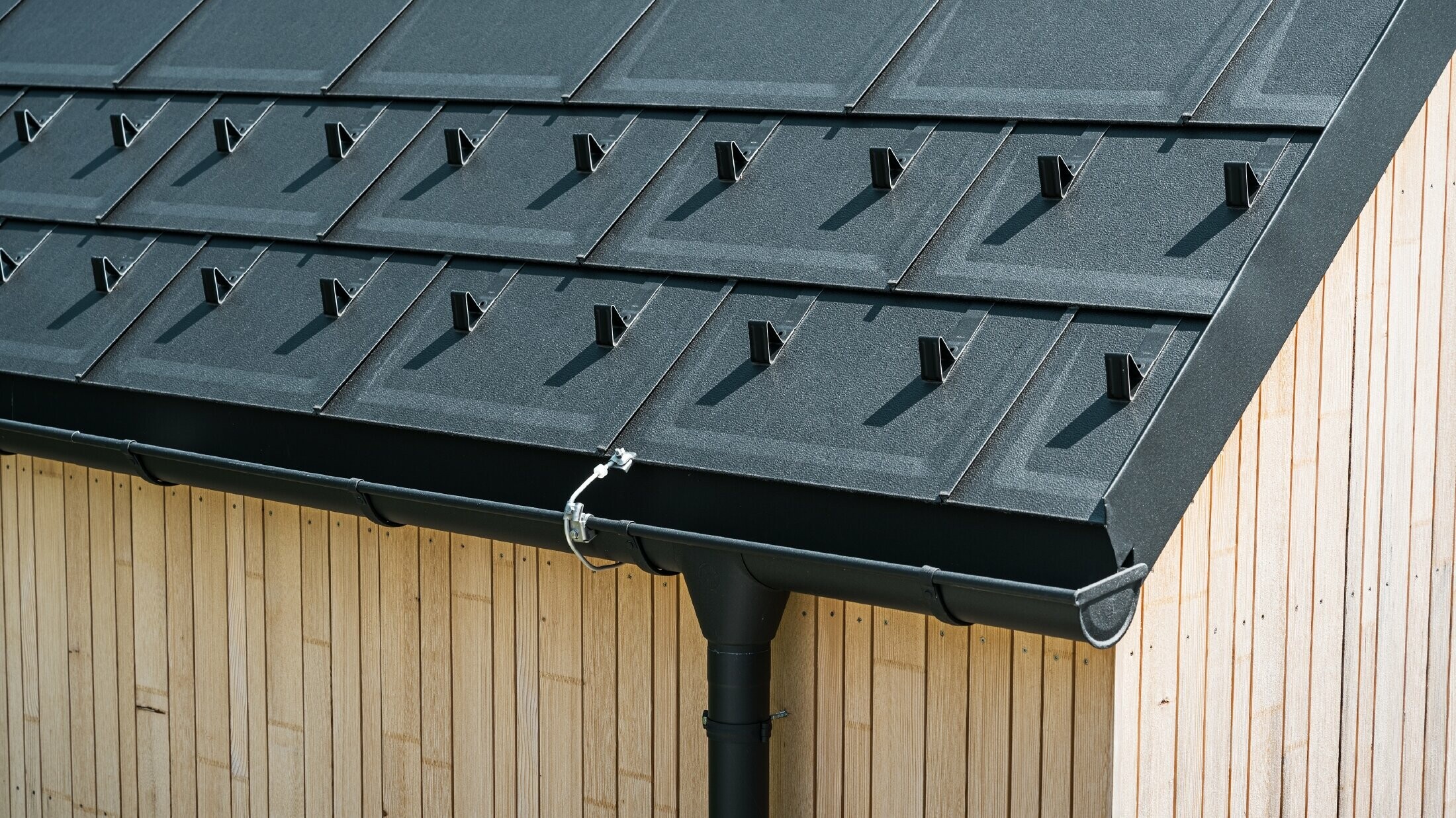 Nahaufnahme der PREFA Dachplatte R.16 mit Schneestoppern, Dachrinne und Ablaufrohr in Schwarz P.10 inkl. Blitzschutz und einer senkrechten Holzfassade