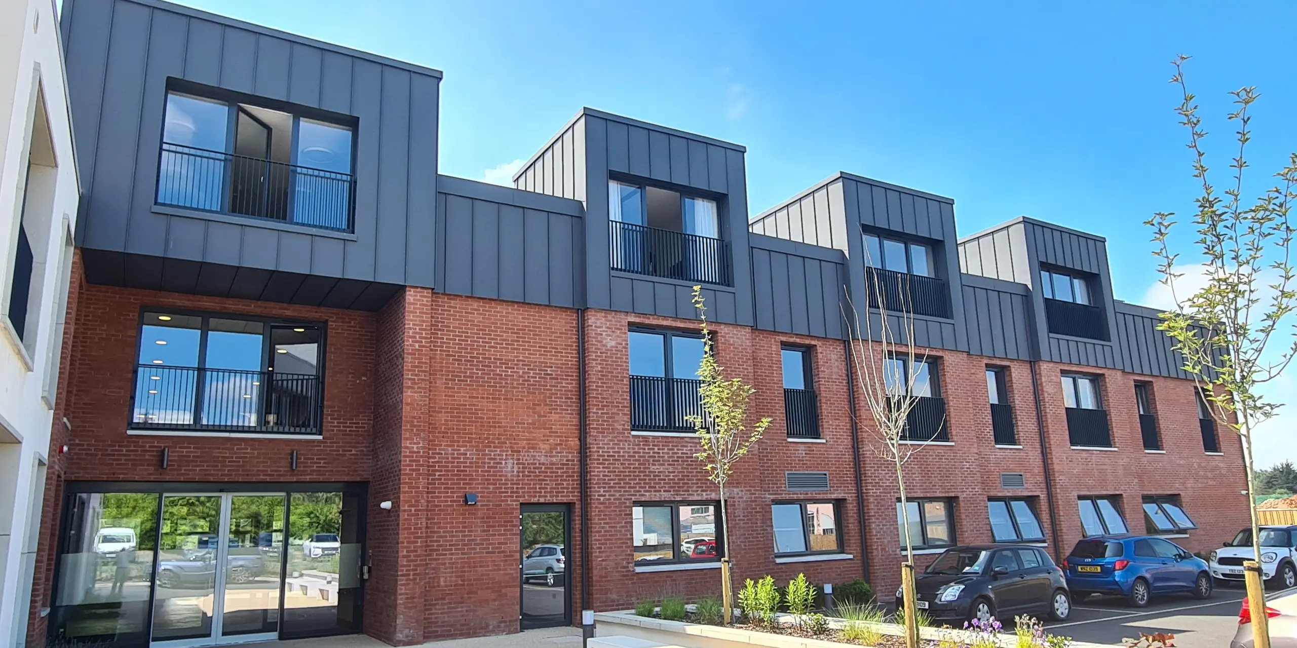 Vorderansicht der Harberton Hall in Belfast, Nordirland. Die Stehfalzdeckung aus PREFALZ P.10 dunkelgrau ergänzt die Ziegelfassade perfekt. Große Fenster und Flachdachgauben unterstreichen das innovative Design des Gebäudes für betreutes Wohnen und schaffen eine einladende Atmosphäre. Die gläserne Eingangstür gewährt Einblicke in das Innere der Einrichtung.