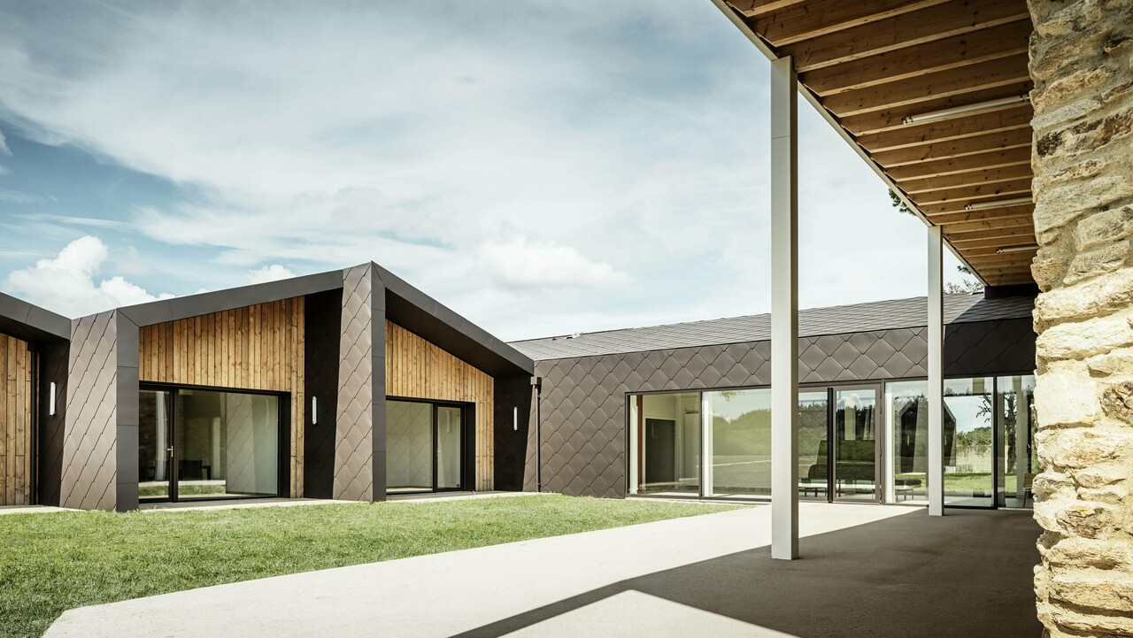 Das Bild zeigt ein modernes Gebäude mit einer Fassade aus PREFA 44 × 44-Dach- und Wandelementen in Walnussbraun. Die Fassade besticht durch ihre elegante Rautenstruktur, die sich harmonisch in die warmen Holzelemente einfügt. Große Glasflächen lassen viel Tageslicht ins Innere und schaffen eine offene und luftige Atmosphäre. Ein überdachter Durchgang auf der rechten Bildseite führt zu einer rustikalen Steinmauer, die einen interessanten Kontrast zum modernen Design bildet.