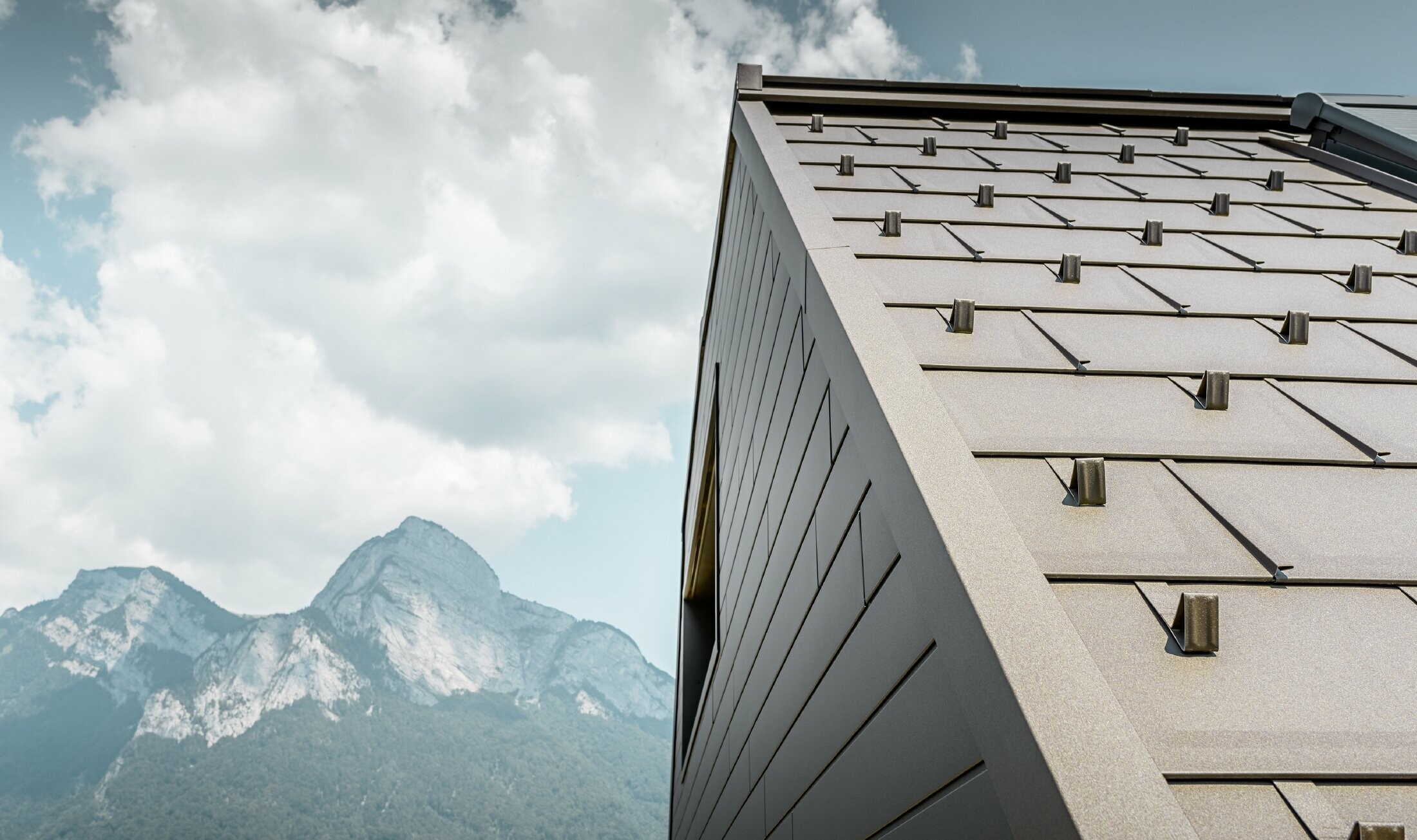 Nahaufnahme der PREFA Dachplatte R.16 in P.10 Braun inkl. Einfassung und Bergen sowie Wolkenhimmel im Hintergrund.