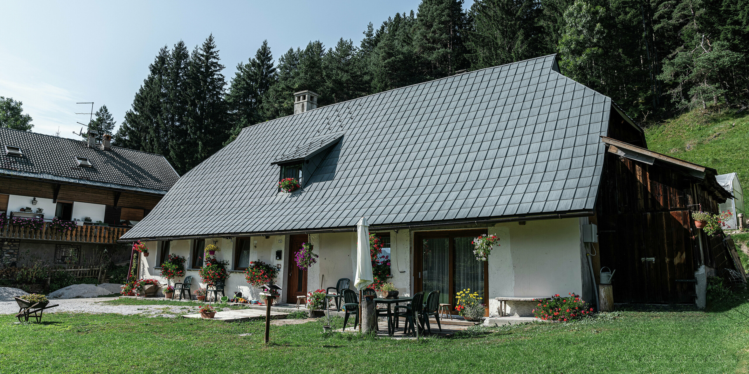 Ein charmantes Einfamilienhaus in Tarvis, Italien, seit 1972 mit langlebigen PREFA Dachplatten eingedeckt: Das große Krüppelwalmdach mit der widerstandsfähigen Aluminiumeindeckung schützt das Haus seit Jahrzehnten zuverlässig vor den alpinen Witterungseinflüssen. Blühende Blumenkästen zieren die rustikale Fassade, eine gemütliche Terrasse mit Gartenmöbeln lädt zum Verweilen ein. Im Hintergrund sorgen dichte, grüne Nadelbäume für eine idyllische Atmosphäre und fügen das Haus perfekt in die natürliche Umgebung ein.