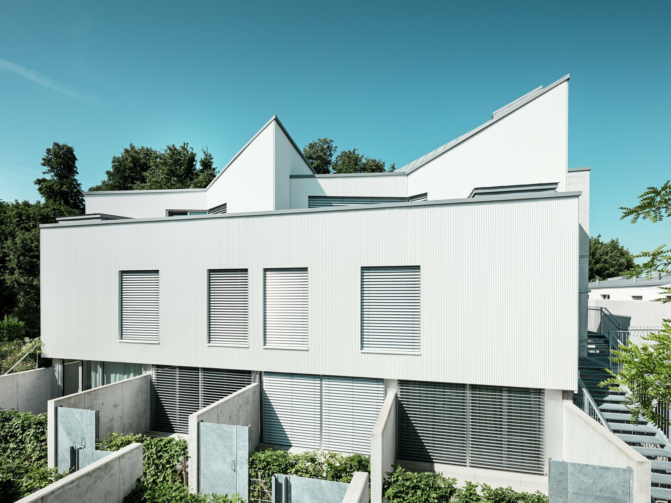 Die zeitgemäßen Tiny Homes am Zollikerberg in der Schweiz überzeugen durch ihre klare Linienführung und die moderne PREFA Aluminiumfassade im Strangpressprofil „Zacke”. Die vertikal verlegten, sanft geschwungenen Profile erzeugen ein markantes und elegantes Fassadenbild, das durch Lichtreflexionen lebendig wirkt. Die weiße Oberfläche sorgt für Leichtigkeit und betont die geometrische Architektur. Die vorgehängte, hinterlüftete Fassade schützt das Gebäude dauerhaft vor Witterungseinflüssen und verbessert das Raumklima. Sie ist pflegeleicht und korrosionsbeständig.