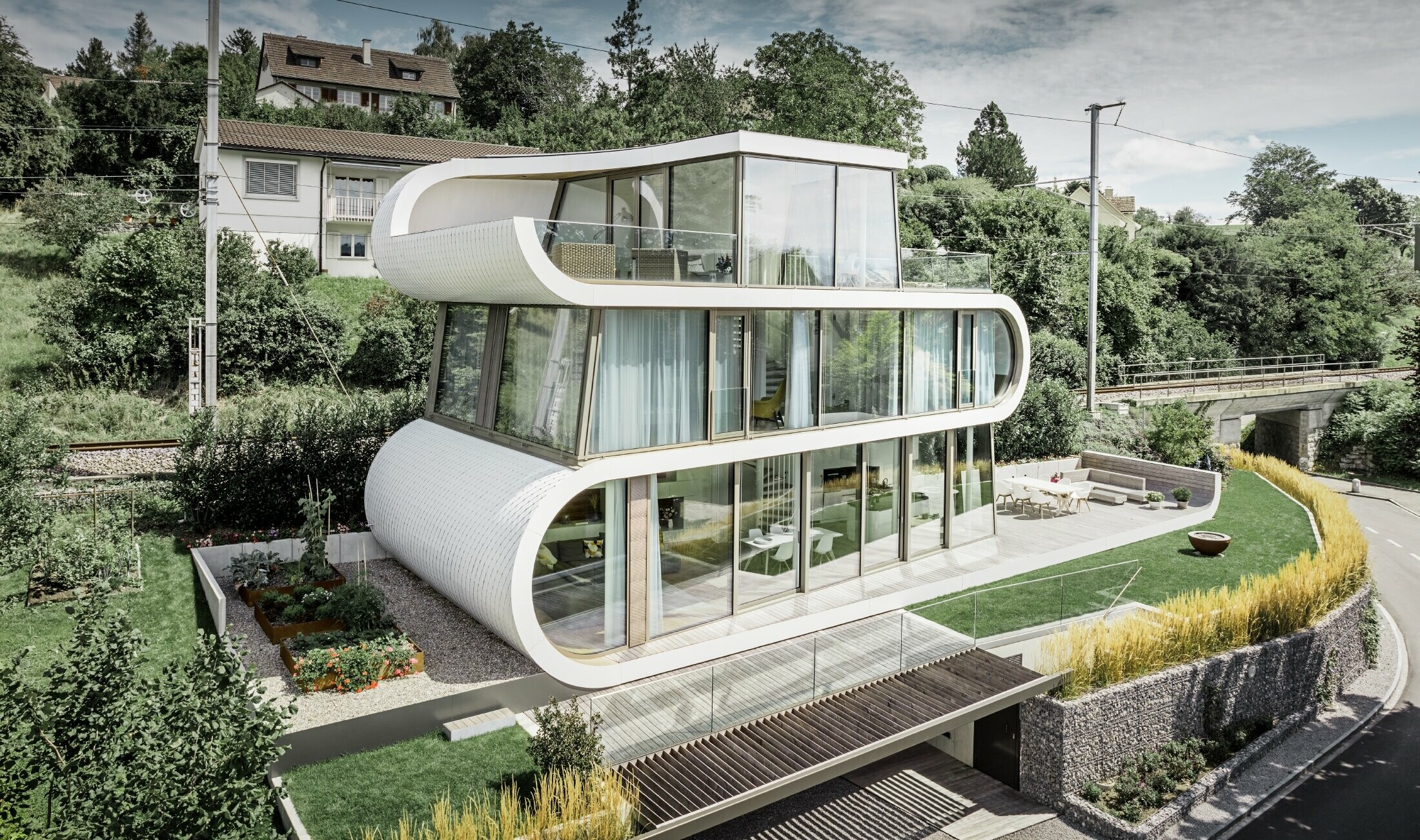 Sehr modernes Einfamilienhaus entworfen durch Architekt Camenzind aus Zürich; Ein geschwungenes Band verbindet die einzelnen Ebenen miteinander. Die Rundungen wurden mit der PREFA Dachraute klein in Reinweiß verkleidet. Das Haus hat viele und große Glasflächen, wodurch das ganze Haus sehr offen wirkt.