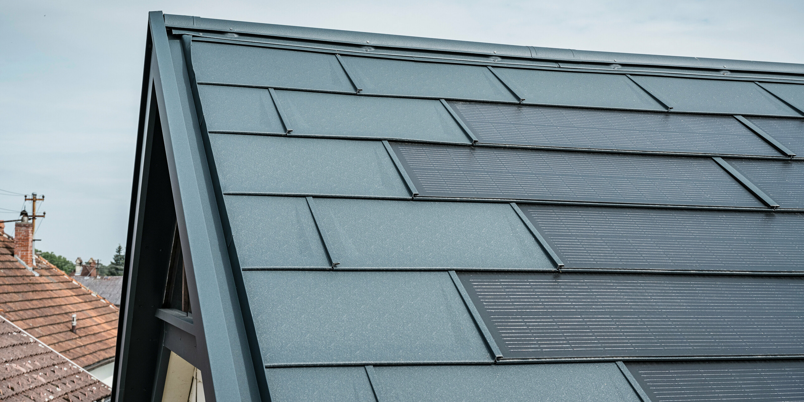 Detailansicht eines Hausdachs ausgestattet mit der innovativen PREFA Solardachplatte. Die Dachplatten mit integrierten Photovoltaikzellen präsentieren sich in einem eleganten Anthrazit. Die homogene Oberfläche fügt sich nahtlos in das Dach ein und sorgt damit für eine moderne und saubere Optik. Das innovative Dachsystem schafft eine effiziente Energienutzung ohne Beeinträchtigung der Ästhetik.