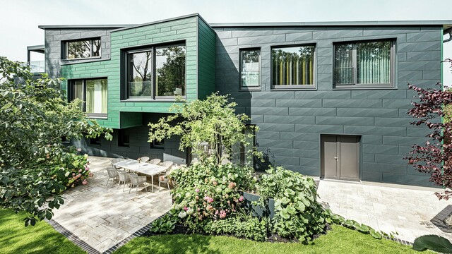 Das Einfamilienhaus in Türkheim beeindruckt durch seine außergewöhnliche Fassade, die mit PREFA Siding X-Paneelen in den Farben Anthrazit und Moosgrün verkleidet ist. Die klaren Linien und die markante Farbwahl der Aluminiumpaneele verleihen dem Gebäude eine moderne und elegante Ausstrahlung. Im Vordergrund ist der großzügige Gartenbereich mit einer stilvollen Terrasse und einem Esstisch mit modernen Stühlen zu sehen. Bunte Blumenbeete und gesunde Pflanzen sorgen für einen lebendigen, grünen Kontrast zur robusten Fassade. Die langlebige und pflegeleichte Aluminiumfassade schützt das Gebäude und sorgt für zeitlose Schönheit.