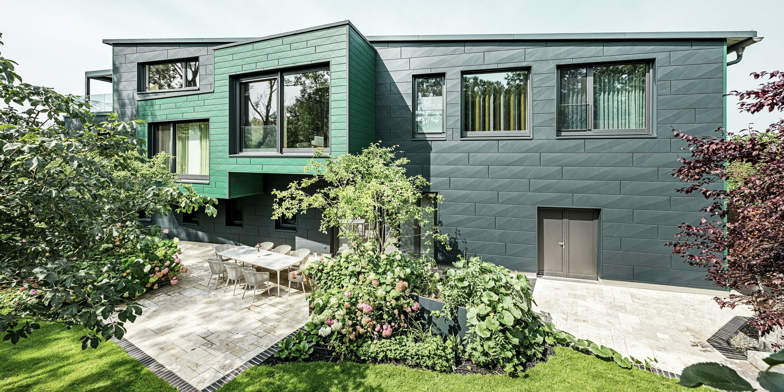 Das Einfamilienhaus in Türkheim beeindruckt durch seine außergewöhnliche Fassade, die mit PREFA Siding X-Paneelen in den Farben Anthrazit und Moosgrün verkleidet ist. Die klaren Linien und die markante Farbwahl der Aluminiumpaneele verleihen dem Gebäude eine moderne und elegante Ausstrahlung. Im Vordergrund ist der großzügige Gartenbereich mit einer stilvollen Terrasse und einem Esstisch mit modernen Stühlen zu sehen. Bunte Blumenbeete und gesunde Pflanzen sorgen für einen lebendigen, grünen Kontrast zur robusten Fassade. Die langlebige und pflegeleichte Aluminiumfassade schützt das Gebäude und sorgt für zeitlose Schönheit.