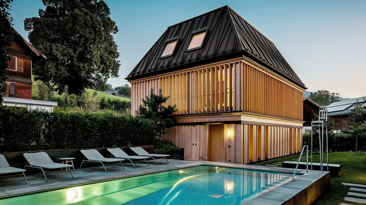 Das „Badehaus“ des Hotel Hirschen in Schwarzenberg in der Dämmerung, mit Blick auf den beleuchteten Außenpool im Vordergrund. Das Gebäude beeindruckt durch sein modernes Walmdach aus PREFALZ in der Farbe P.10 Nussbraun mit präzise ausgeführten, teils schräg verlaufenden Stehfalzen. Zwei Dachfenster sorgen für Tageslicht im Inneren. Die Fassade besteht aus vertikalen, geschoßhohen Holzlamellen, die dem Baukörper Struktur und Leichtigkeit verleihen. Im unteren Bereich dominiert naturbelassenes Holz. Umgeben von gepflegtem Grün und eingebettet in die Hügellandschaft des Bregenzerwaldes entsteht eine harmonische Verbindung aus Architektur, Natur und Erholung.