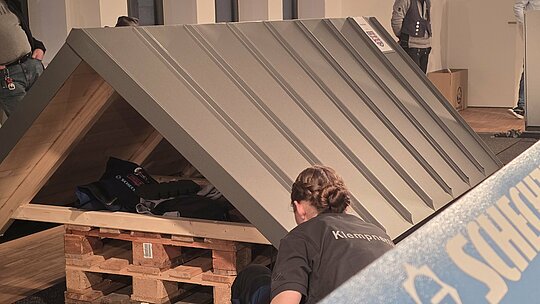 Eine Teilnehmerin arbeitet an einem Dachmodell, das auf einer Holzpalette steht. Das Blechdach ist bereits verlegt, und die Person überprüft die letzte Details, um eine perfekte Ausführung zu erreichen. Im Hintergrund sind andere Teilnehmer bei der Arbeit zu sehen.