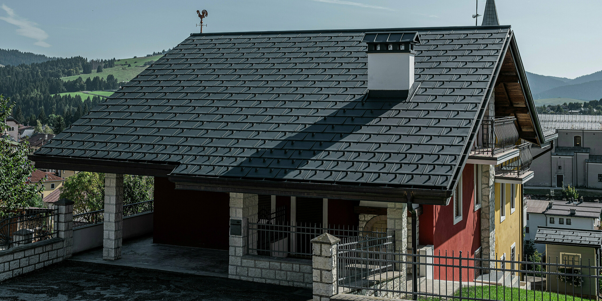 Detailansicht des Eingangsbereiches eines Einfamilienhauses in Gallio mit einem robusten PREFA Dach aus Dachplatten R.16 in P.10 Dunkelgrau. Die Dacheindeckung aus Aluminium sorgt für eine edle Optik und bietet gleichzeitig höchsten Schutz vor Witterungseinflüssen. Die präzise Verarbeitung und das langlebige Material garantieren eine wartungsfreie Lösung für Jahrzehnte. Der massive Kamin mit Natursteinverkleidung und die traditionelle Wetterfahne setzen stilvolle Akzente. Eine perfekte Kombination aus moderner Technik und alpinem Charme.