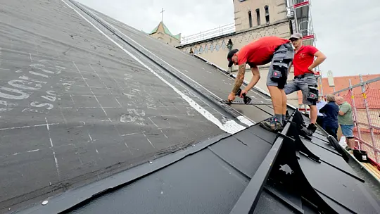 Das Bild zeigt die Verlegung von dachintegrierten Photovoltaikanlagen auf dem Dach einer Kirche in Bayern. Im Vordergrund sind die Solardachplatten deutlich zu erkennen, die ordentlich in Reihen verlegt werden. Die Platten haben eine dunkle, glänzende Oberfläche. Im Hintergrund sieht man einen Teil des Kirchendachs sowie die charakteristischen Merkmale der Kirche, z.B. ein Turm und ein Kreuz. Zwei Arbeiter sind auf dem Dach beschäftigt: Einer führt gerade Montagetätigkeiten aus, während der andere die Platten überprüft. 