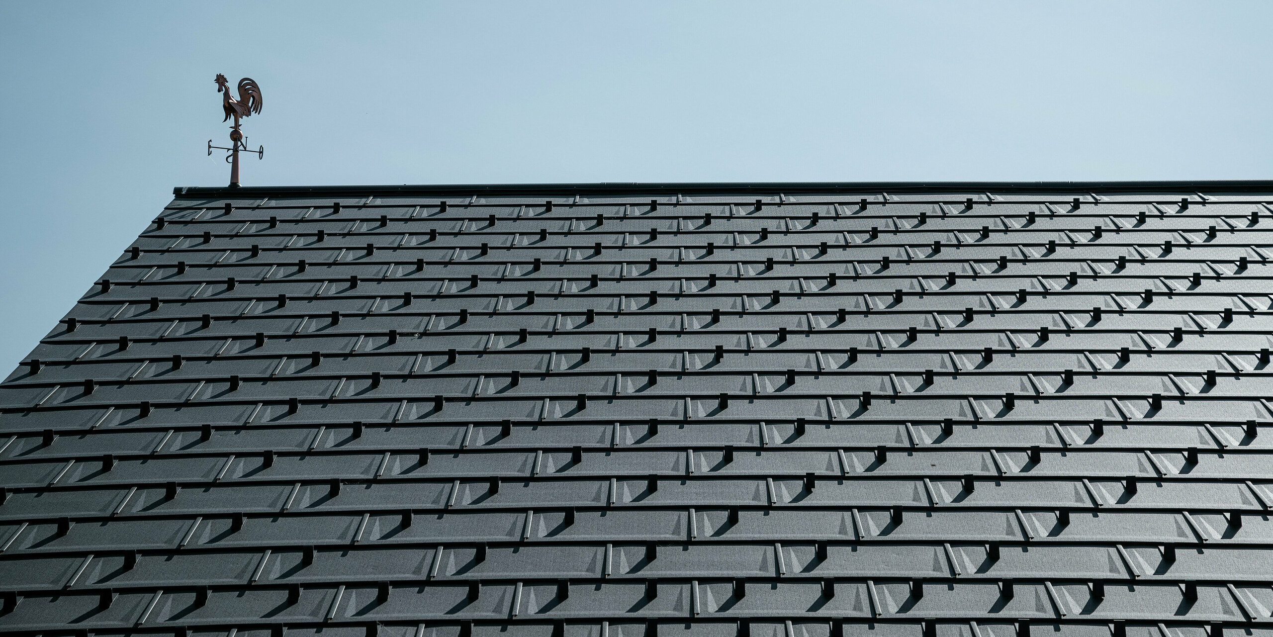 Detailansicht eines PREFA Daches mit den Dachplatten R.16 in P.10 Funkelgrau. Die klare, moderne Optik der Aluminium-Dachplatten sorgt für eine langlebige, witterungsbeständige und wartungsfreie Dachlösung. Die Schneestopper bieten zusätzliche Sicherheit in schneereichen Regionen. Gekrönt wird das Dach von einer klassischen Wetterfahne, die Tradition mit moderner Technik verbindet. Dank der robusten PREFA Dachplatten R.16 bleibt das Dach über Jahrzehnte stabil und ästhetisch.