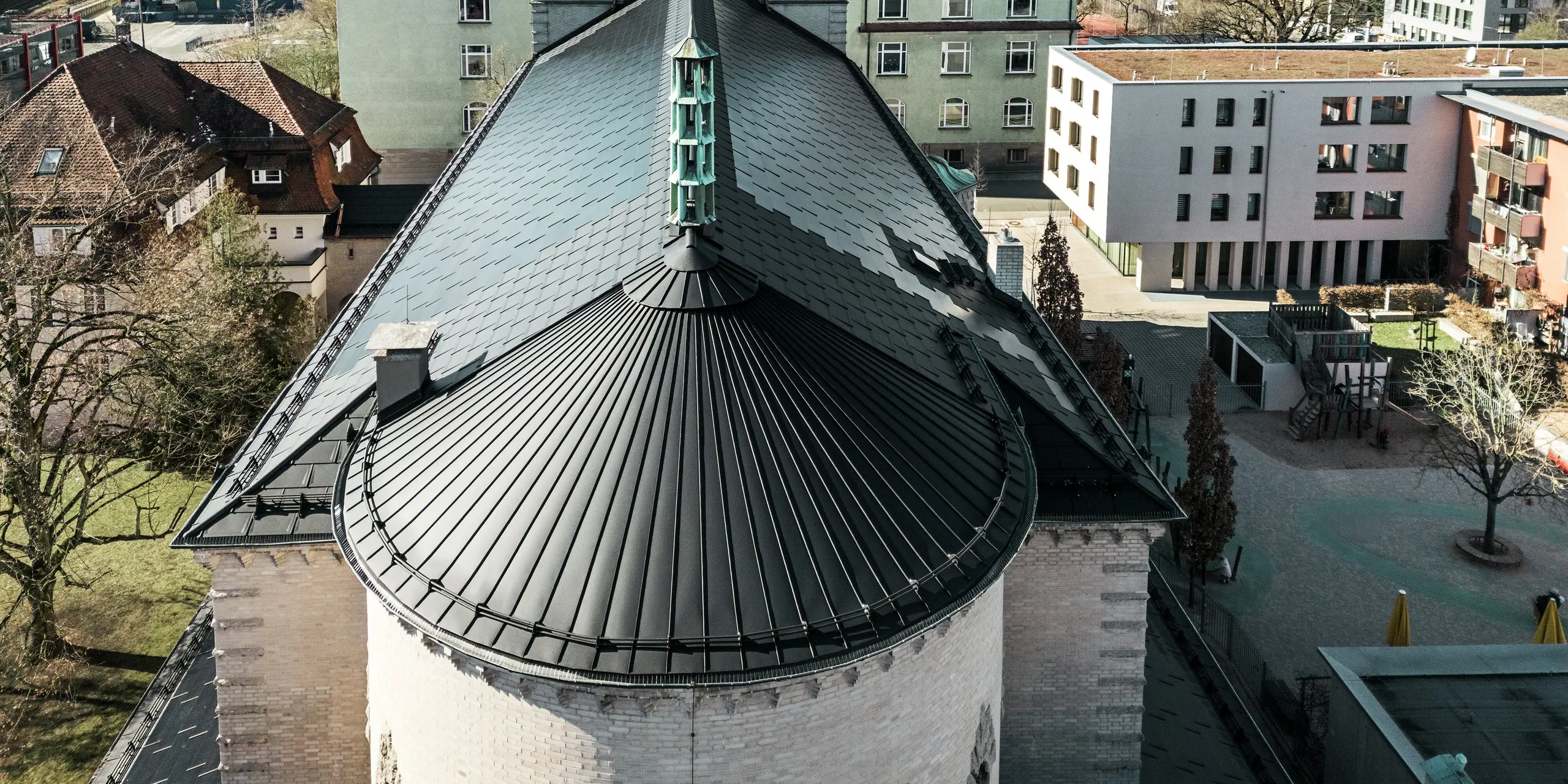 Das Bild zeigt das markante, rund gewölbte Dach auf der Rückseite der St. Karl Borromäus Kirche in Nürnberg. Die Gebäuderundung wurde mit einer Stehfalzdeckung aus schwarzem Aluminium eingedeckt. Das Satteldach, das den Hauptteil des Gebäudes schützt, wurde mit schwarzen PREFA Solardachplatten eingedeckt. Die innovativen Solardachplatten fungieren als optisches Highlight und liefern gleichzeitig Strom aus erneuerbarer Energie. Der Blick auf das Dach und die zentral positionierte Turmspitze verdeutlicht die harmonische Integration der modernen Solartechnologie in die historische Architektur. Im Hintergrund sind sowohl traditionelle als auch moderne Gebäude zu sehen, was die gelungene Verbindung von Denkmalschutz und nachhaltigem Bauen unterstreicht.
