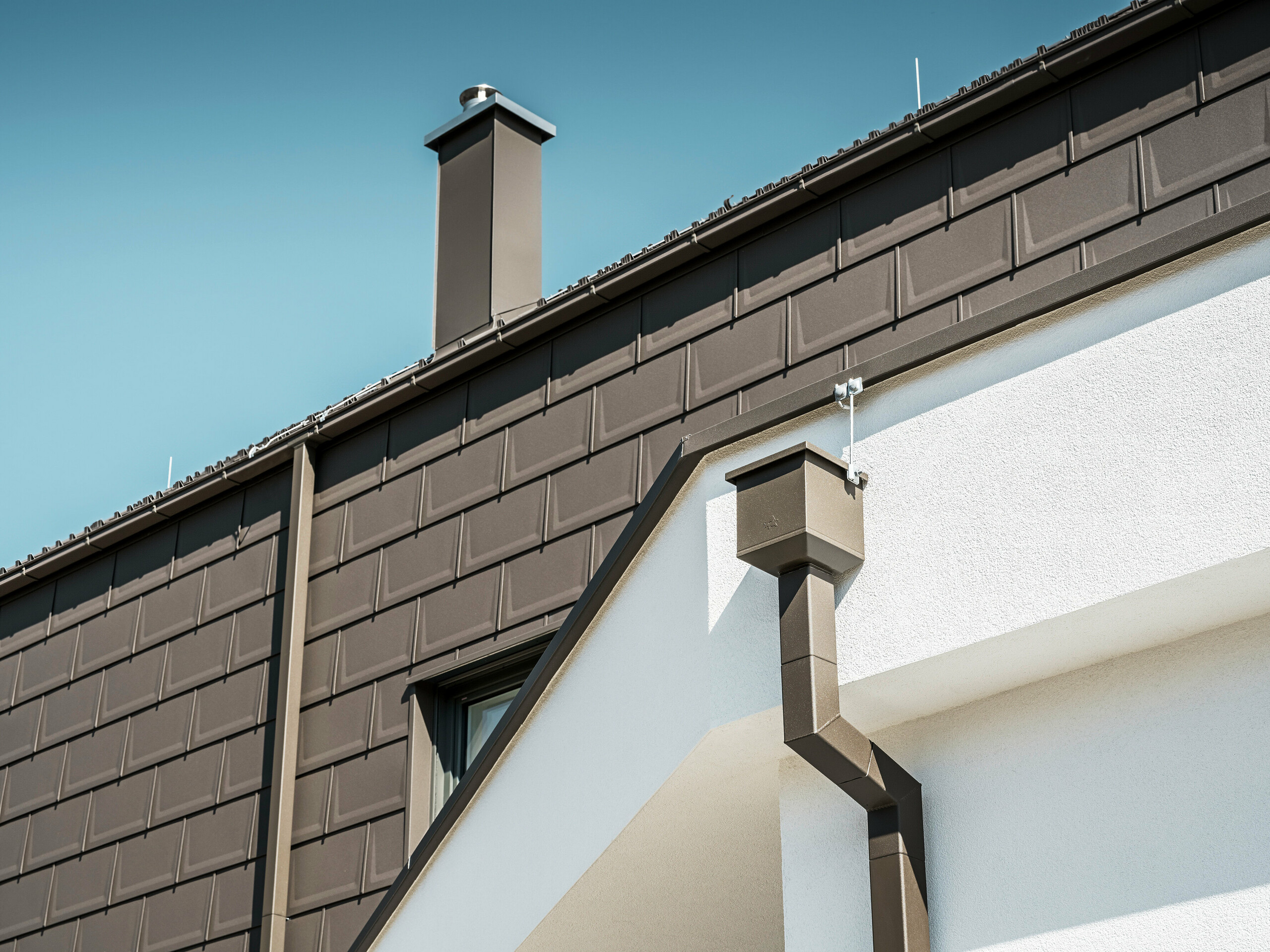 Detailaufnahme des Dachrandes bzw. der Fassade eines Hauses in Neukirchen, Österreich, ausgestattet mit PREFA R.16 Dachplatten in P.10 braun. Sowohl die Dachplatten, die bei diesem Gebäude als Dach- und Fassadenverkleidung dienen, als auch die Dachentwässerung, bestehend aus Vierkantrohr, Wassersammelkasten und Kastenrinne, sind in der gleichen Farbe gehalten und bieten eine einheitliche, hochwertige Ausführung. Die Verwendung von PREFA Produkten verleiht dem Gebäude ein zeitgemäßes Aussehen, während die ästhetische Einbindung der Entwässerungselemente die funktionale Eleganz des Designs unterstreicht.