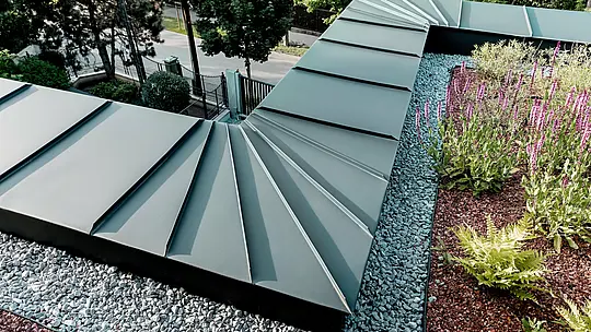 Die Aufnahme zeigt eine detaillierte Nahansicht der Attika‑Ausführung der Budapester Mehrgenerationen‑Villa, verkleidet mit PREFALZ Aluminium in P.10 Anthrazit. Die fächerförmig angeordneten Scharen laufen präzise an der Gebäudekante zusammen und demonstrieren die hohe Formbarkeit und Verarbeitungsqualität der PREFA Aluminiumprodukte. Das anthrazitfarbene Metall bildet einen eleganten Kontrast zum begrünten Dachbereich mit Kiesbett und blühenden Pflanzen. Die Szene betont die Langlebigkeit, Witterungsbeständigkeit und technische Raffinesse der PREFA Lösung.
