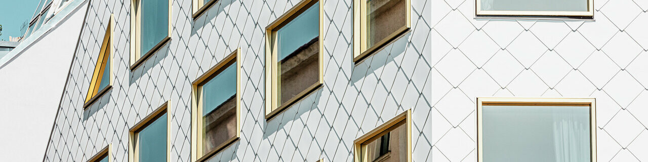 Ein modernes Viersternehotel in Wien, verkleidet mit PREFA Dach- und Wandrauten 44 × 44 in P.10 Reinweiß. Die Fassade zeigt eine blockhafte Struktur mit einer Vielzahl von spiegelverglasten Fenstern, die in einer scharfen Rasterung angeordnet sind und die Bewegungen der Umgebung reflektieren.