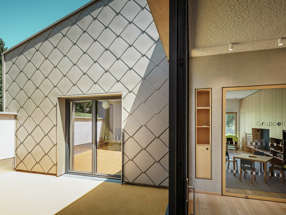 Modernes Gebäude mit PREFA Dach- und Fassadenverkleidung aus 44 x 44 Rautenelementen, fotografiert bei Tageslicht. Das Gebäude zeigt eine harmonische Integration traditioneller und moderner architektonischer Elemente, mit den Metallrauten als markanten Elementen. Die großen Fenster geben den Blick frei auf einen hellen Innenraum, den so genannten Gruppenraum, der mit Tischen und Stühlen für Besprechungen oder Unterricht ausgestattet ist. Übersetzt mit DeepL.com (kostenlose Version)