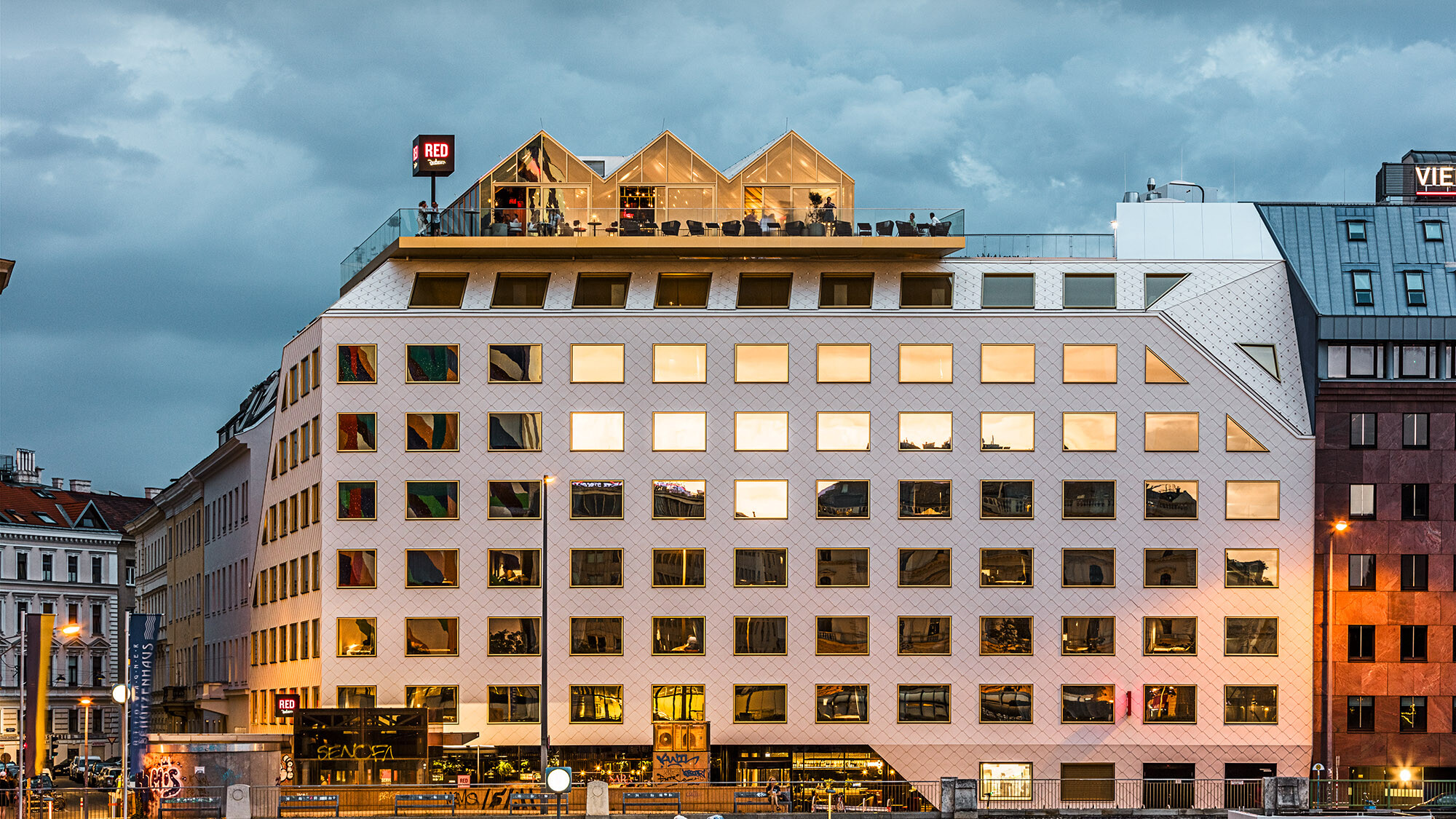 Eine Abendaufnahme des Hotels THE ROCK Radisson RED Vienna samt seiner belebten Umgebung; die gegenüberliegenden Gebäude spiegeln sich in seinen Fenstern.