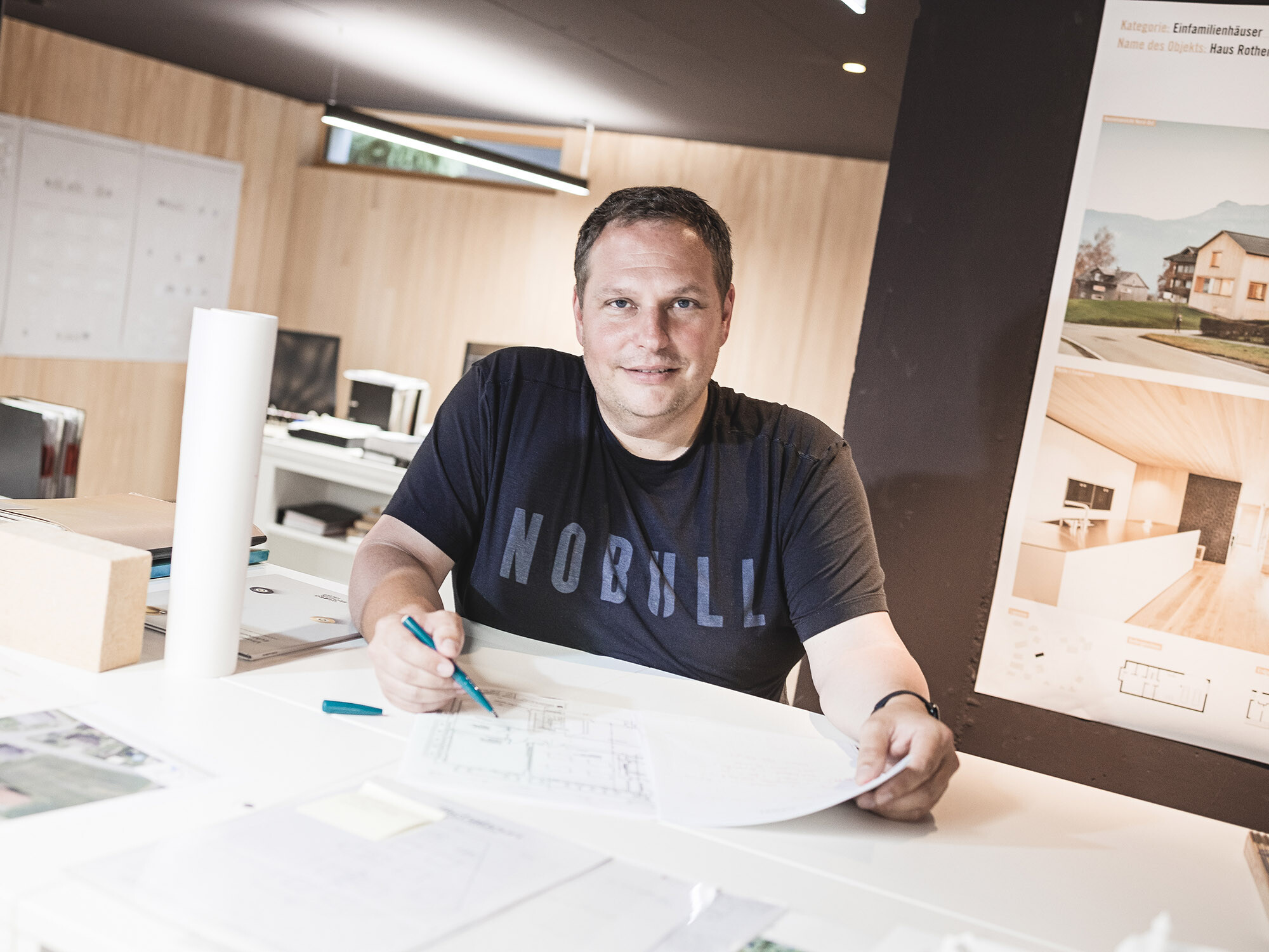 Portrait des Baumeisters Jürgen Haller an seinem Büroschreibtisch, einen Stift in der einen Hand, einen Plan in der anderen Hand haltend.