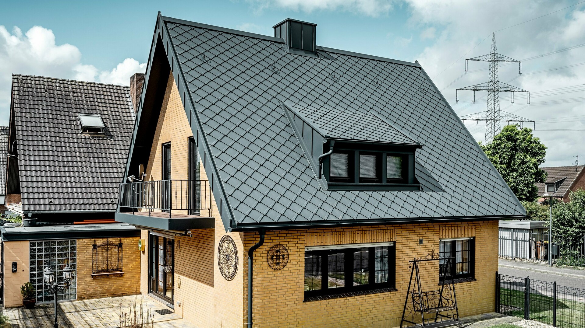 Einfamilienhaus aus gelbem Klinker nach der Dachsanierung mit PREFA Dachraute 29 × 29 in Dunkelgrau; neu eingedecktes, steiles Satteldach mit Rautenmuster, integrierter Gaube und Kamin, Terrasse und Garten im Vordergrund.