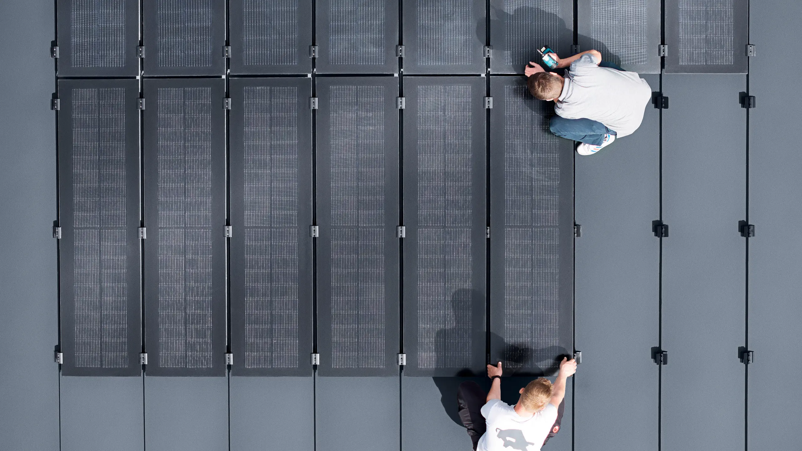 Zwei Personen installieren PREFALZ-Solarmodule auf einer großen grauen Dachfläche, die mit doppelflanschigen Paneelen bedeckt ist.