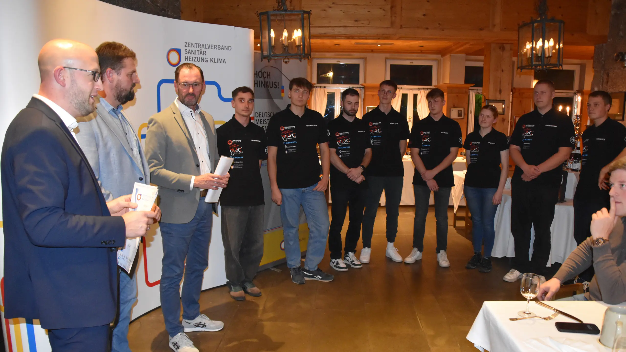 Die Gewinner und Teilnehmer der Meisterschaft stehen aufgereiht, während sie die Urkunden und Preise entgegennehmen. Im Vordergrund sprechen die Vertreter des ZVSHK und der PREFA, während die Auszubildenden aufmerksam zuhören.