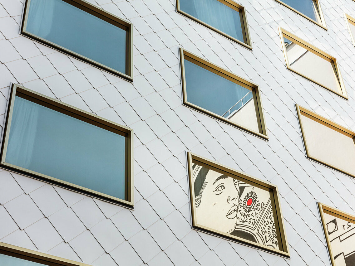 Detailaufnahme der Fassade des "THE ROCK Radisson RED Vienna" Hotels, das mit PREFA Dach- und Wandrauten 44 × 44 in P.10 Reinweiß verkleidet ist. Unter den mehreren spiegelverglasten Fenstern mit goldenen Rahmen sticht ein Fenster hervor, in das ein Kunstwerk eingelassen ist, welches das Porträt einer Person mit einem markanten roten Akzent darstellt