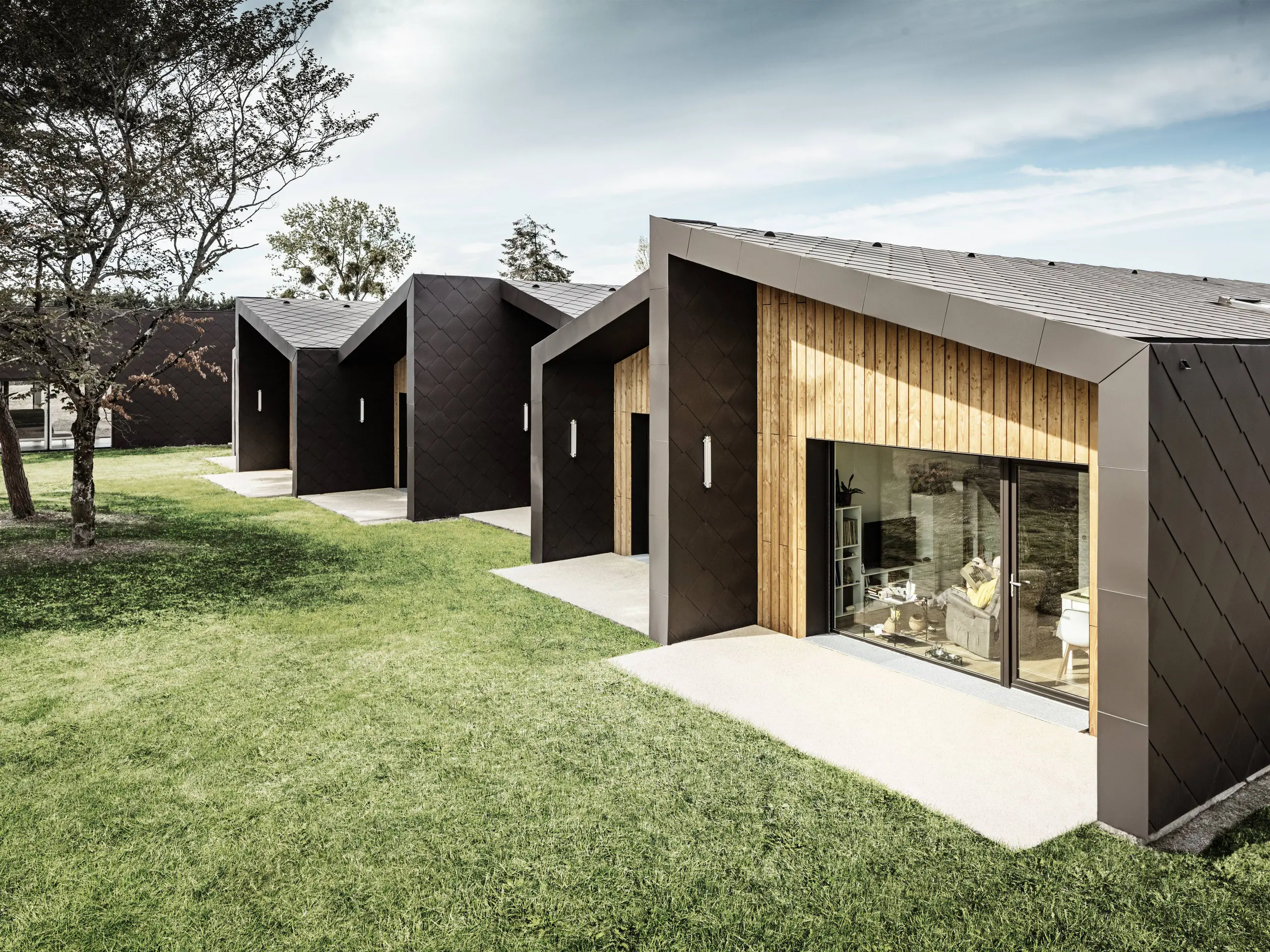 Das Bild zeigt eine Reihe moderner, minimalistisch gestalteter Gebäude mit einer Fassade aus PREFA 44 × 44 Dach- und Wandrauten in Walnussbraun. Die Rautenstruktur der Verkleidung wird harmonisch durch warme Holzelemente ergänzt, die die Eingangsbereiche und großen Glasflächen einrahmen. Die Gebäude befinden sich in einer grünen, sorgfältig gestalteten Landschaft, die eine ruhige und angenehme Atmosphäre schafft. Dieser Entwurf vereint Funktionalität mit ästhetisch ansprechender Architektur.