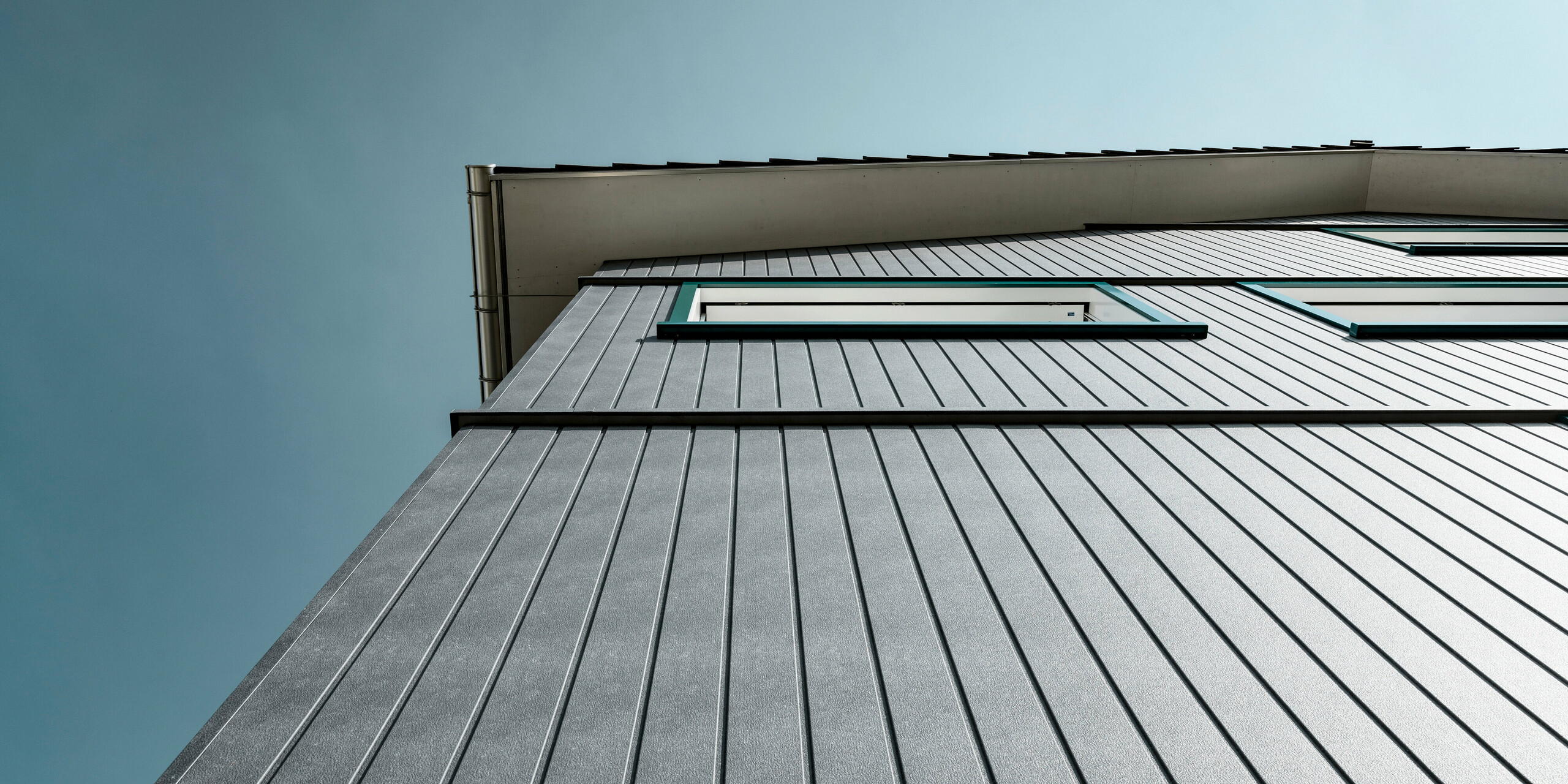 Architektonische Perspektive eines Mehrfamilienhauses in Weissbad, Schweiz, hervorgehoben durch die PREFA Siding Aluminiumfassade in P.10 Steingrau. Die Fassadenpaneele sind vertikal montiert, was dem Gebäude ein gut strukturiertes Erscheinungsbild verleiht. Das Spiel aus Linien und die schlanke Silhouette der Sidings betonen die Höhe des Gebäudes und dessen moderne Architektur unter dem blauen Himmel.