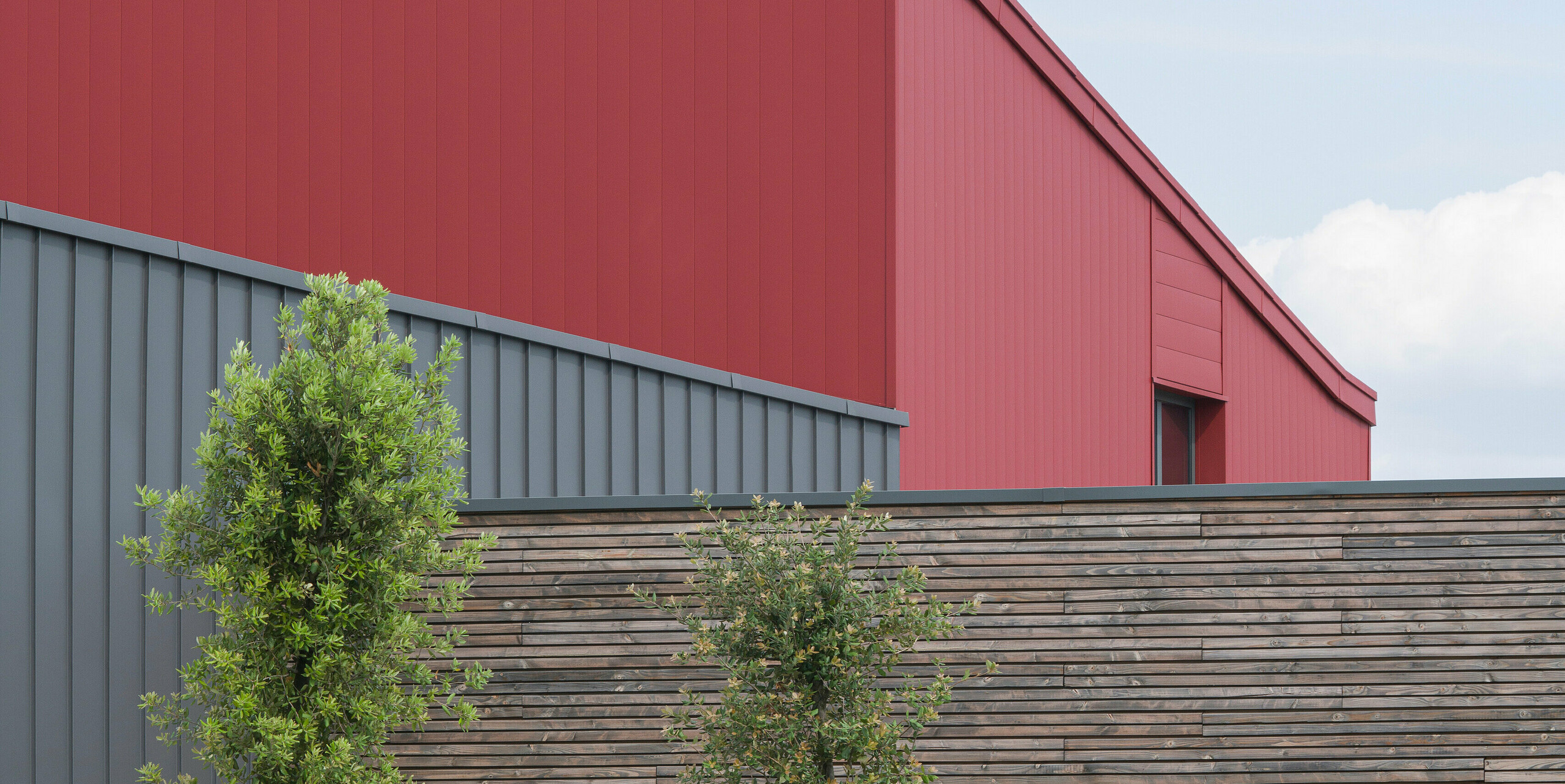Ein modernes Gewerbegebäude mit einer auffälligen roten Aluminiumfassade. Die Farbwahl spricht eine klare architektonische Sprache. Die strukturierte, graue Aluminiumverkleidung bildet einen sanften Kontrast zum kräftigen Rot. Die langlebige Aluminiumverkleidung PREFALZ an der Fassade und am Dach sorgt für idealen Schutz vor jeder Witterung. Die ergänzende Naturholzverkleidung vermittelt Wärme und Natürlichkeit. Vor dem Gebäude sind junge Bäume zu sehen, die sorgfältig mit Stützpfählen befestigt sind. Ein gepflegtes Blumenbeet fügt zusätzliche Farbe und Leben hinzu. Eine Elektrofahrzeug-Ladestation im Vordergrund zeigt das Engagement für nachhaltige Energielösungen.