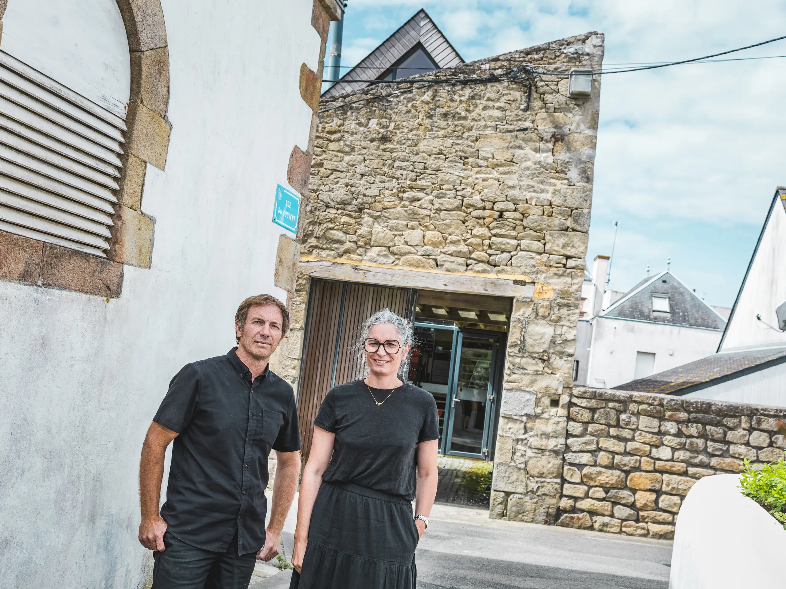 In einer schmalen bretonischen Gasse stehen die beiden Architekten Anne-Charlotte und Jean-François Riguidel vor einer markanten Natursteinfassade. Die beiden sind komplett in Schwarz gekleidet. Der spannende Kontrast aus traditionellem Mauerwerk, weißen Hausfassaden und moderner Dacharchitektur lenkt den Blick auf die gelungene Verbindung von regionaler Baukultur und zeitgemäßem Design.