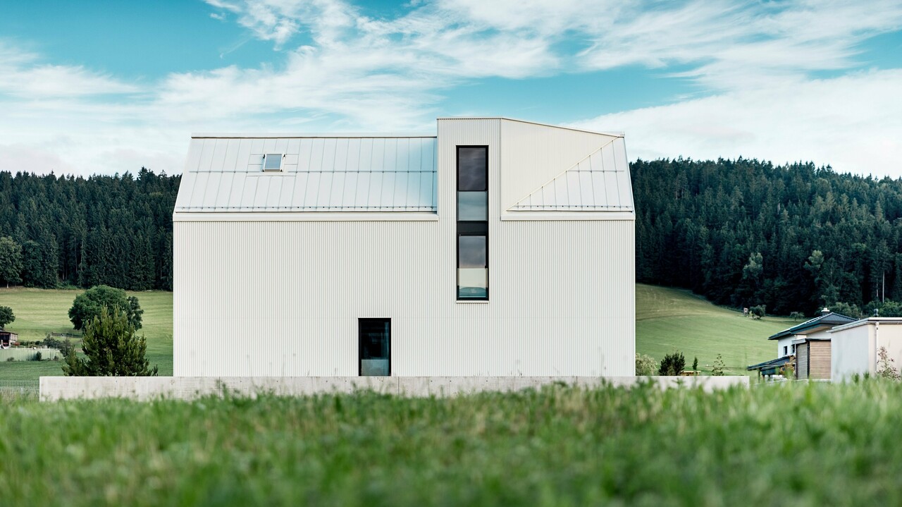 Das Bild zeigt das Einfamilienhaus in Freistadt aus einer Seitenansicht, die die markante, minimalistische Architektur des Gebäudes hervorhebt. Die Fassade aus reinweißem PREFALZ-Aluminium und das steile Satteldach schaffen eine klare, moderne Silhouette, die sich harmonisch in die grüne Umgebung einfügt. Die großen Fensterflächen und das elegante Gaubenfenster betonen das funktionale Design, während das hochwertige Aluminiummaterial sowohl ästhetische Beständigkeit als auch Witterungsresistenz gewährleistet. Das Gebäude strahlt durch seine einfache Form und die Verwendung langlebiger Materialien eine zeitlose Eleganz aus und stellt eine perfekte Kombination aus Architektur und Naturverbundenheit dar.