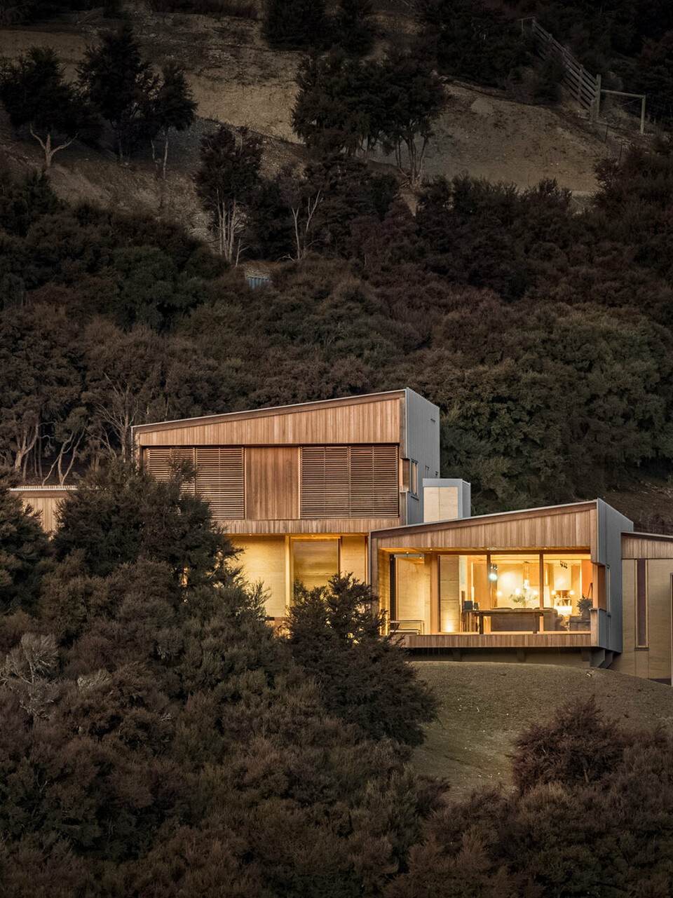 Abendaufnahme des von innen beleuchteten Einfamilienhauses, eingebettet in die Hügellandschaft rund um Wanaka, Neuseeland. Ein Teil des Gebäudes wird vom Buschwald verdeckt.