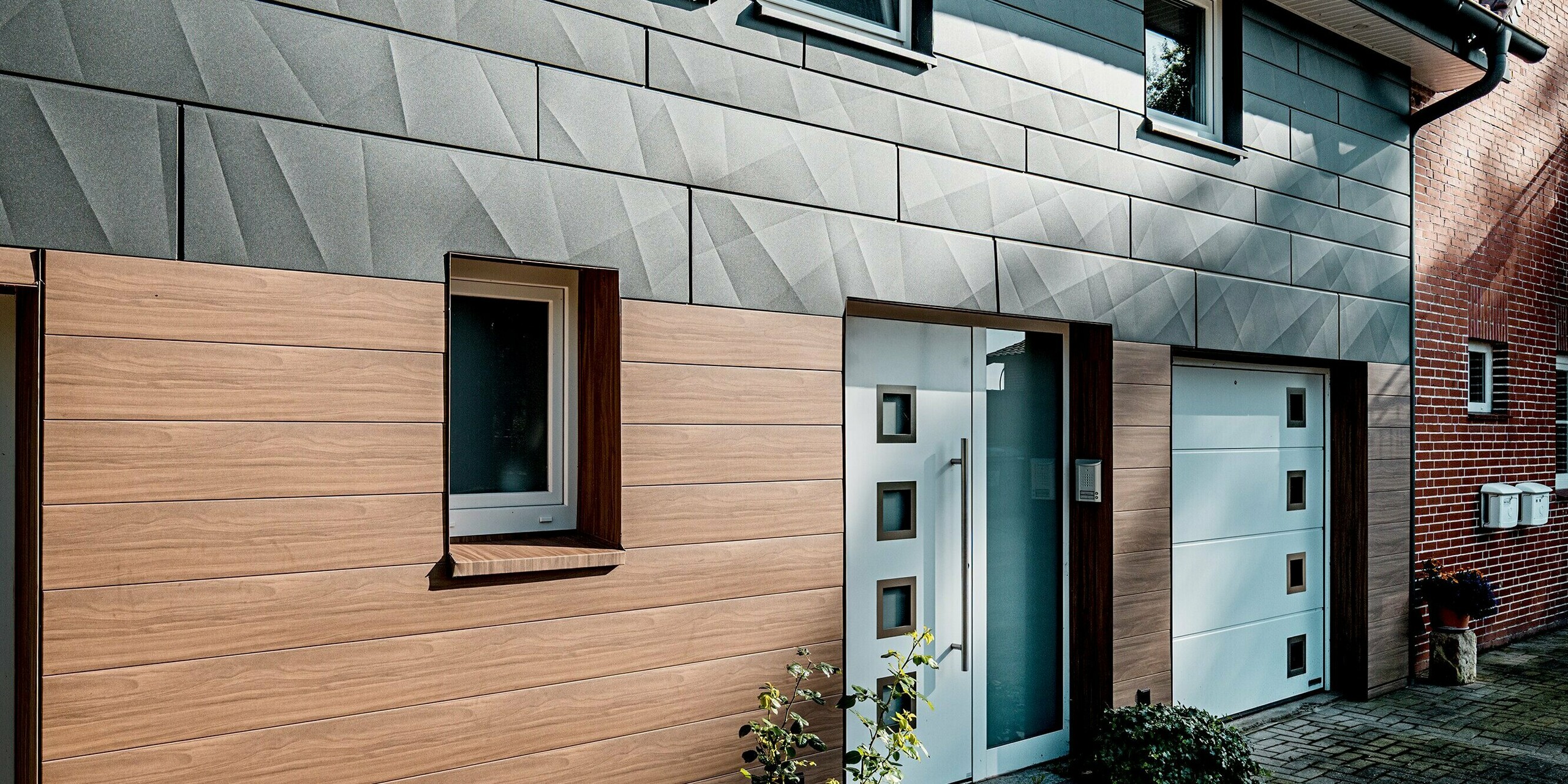 Detailansicht des Eingangsbereichs eines sanierten Einfamilienhauses mit moderner PREFA Aluminiumfassade. Im Vordergrund überzeugt PREFA Siding in Walnussbraun durch seine hochwertige Holzoptik, die dem Sockel- und Eingangsbereich Wärme und Wohnlichkeit verleiht. Der Hauseingang mit weißer Tür und klaren Glaselementen ist präzise eingefasst und setzt einen eleganten Akzent. Darüber sorgt PREFA Siding.X in P.10 Dunkelgrau mit markanten Kantungen für eine dynamische Fassadenstruktur und spannende Licht- und Schattenspiele. Die langlebigen, pflegeleichten und witterungsbeständigen Aluminiumpaneele verbinden zeitgemäßes Design mit nachhaltiger Funktionalität und hoher architektonischer Qualität.