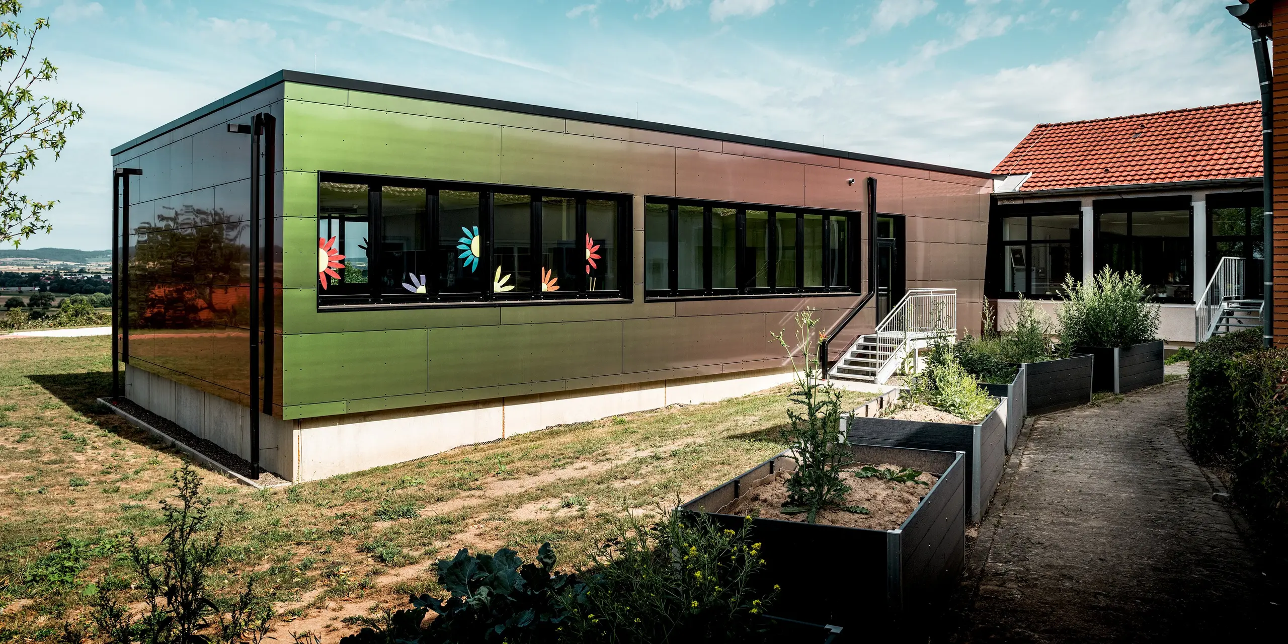 Farbenfroher Schulanbau der Johannwolfschule Außenstelle Angerstein: Die schimmernde PREFA Fassade aus Aluminium Verbundplatten in der Sonderfarbe Spring Lake begeistert mit wechselnden Grün-, Bronze- und Rottönen. Große Fenster mit bunten Blumenmotiven, eine Außentreppe und bepflanzte Hochbeete prägen die einladende Szene. Das dunkle PREFA Quadratrohr ergänzt die langlebige, witterungsbeständige Aluminiumfassade.