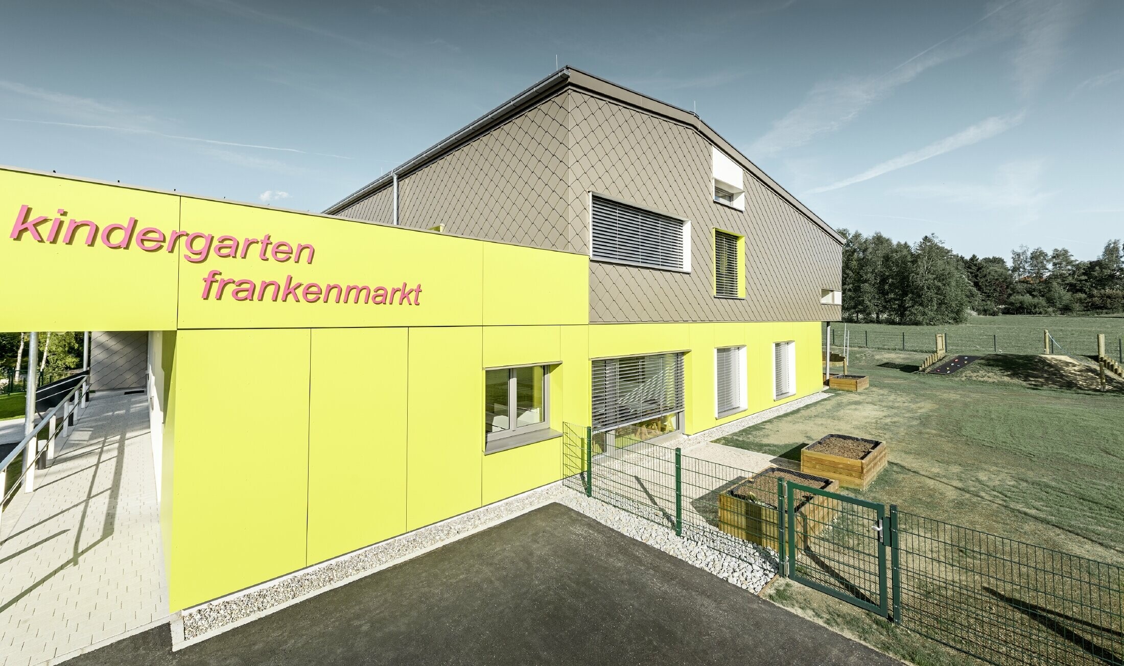 Die Fassade des Kindergarten in Frankenmarkt wurde zum Teil mit der PREFA Wandraute in Bronze verkleidet. Der Rest der Fassade wurde mit Fassadenplatten in Gelb bekleidet. Man erkennt auch den Schriftzug des Kindergartens.
