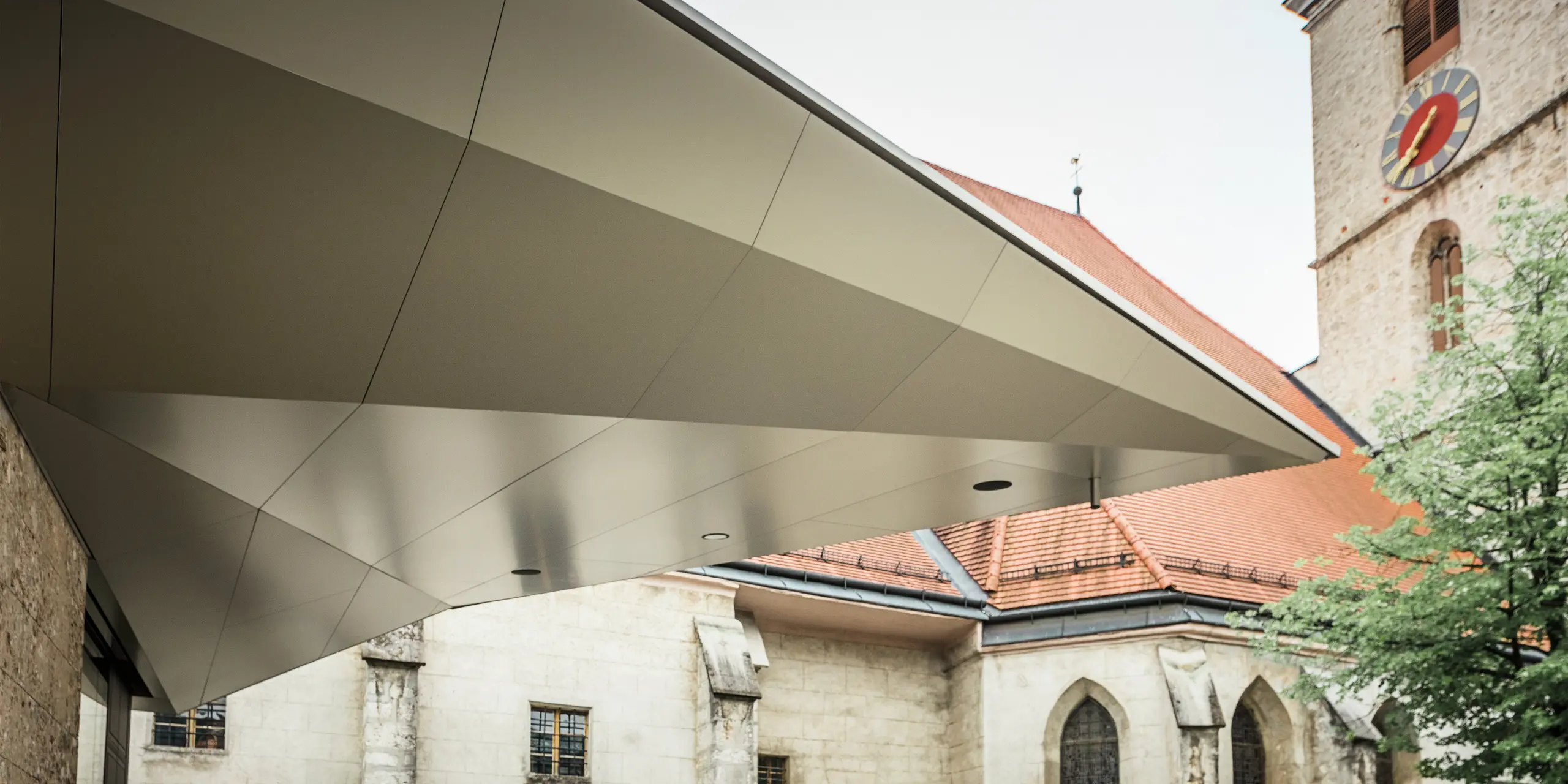 Dynamisch gespannte Dachfläche mit scharfkantiger Linienführung: PREFA Aluminium Verbundplatten ermöglichen diese präzise, fugenarme Gestaltung, die dem Eingangsbereich Leichtigkeit und Modernität verleiht - bei gleichzeitig hoher Witterungsbeständigkeit. Im Hintergrund ist die historische Kirche aus dem 14. Jahrhundert zu erkennen.