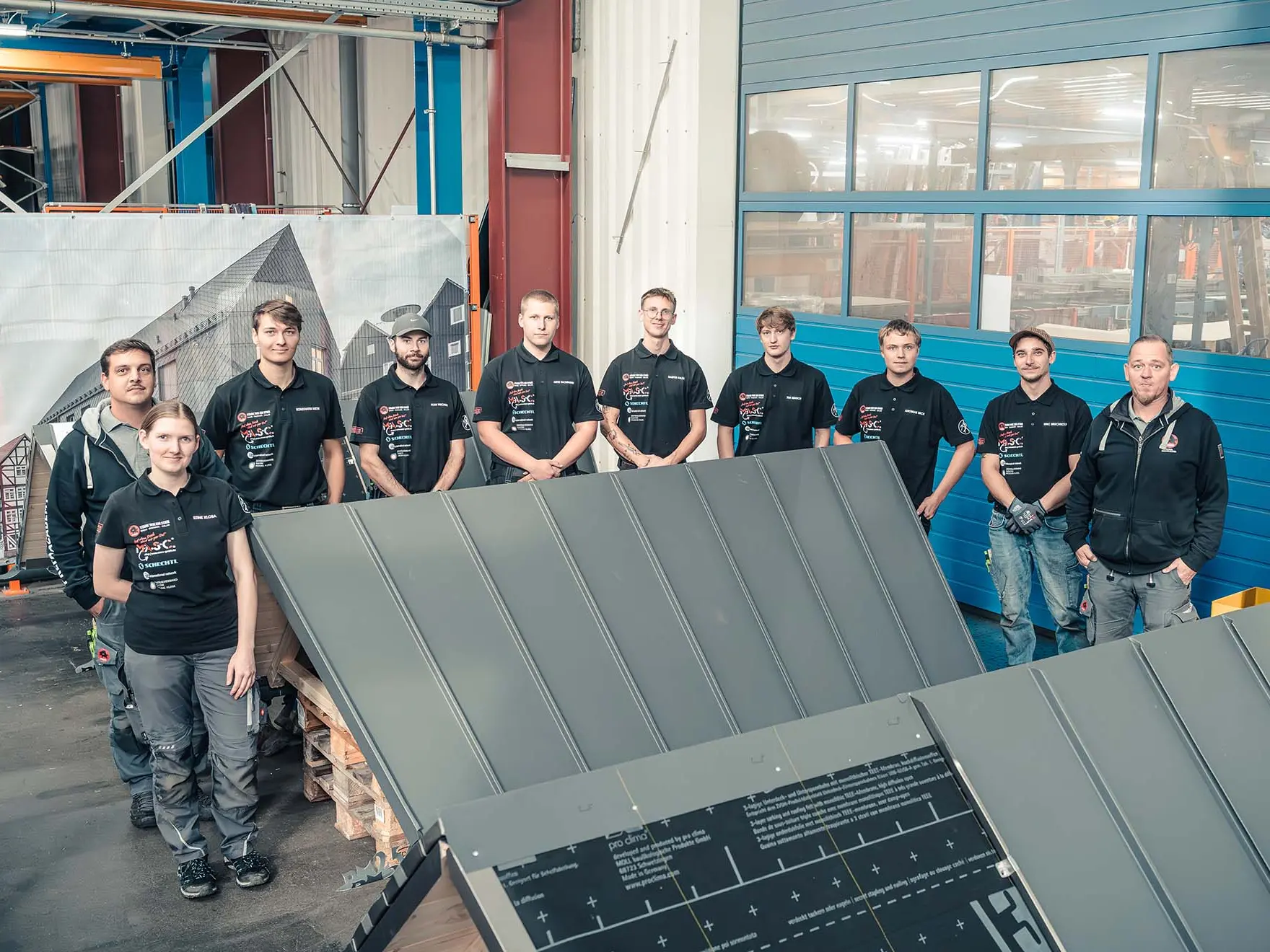 Gruppenfoto der acht teilnehmenden Meisterschüler der diesjährigen Deutschen Meisterschaft im Klempnerhandwerk in der PREFA Academy in Wasungen. Mehrere Teilnehmende stehen in der Werkhalle hinter Übungsmodellen aus Aluminium mit Stehfalztechnik. Alle tragen schwarze PREFA-Trainingsshirts mit den Sponsorenlogos. Mit im Bild sind die Trainer Oliver Kroll und Thomas Schilke, die das Training beaufsichtigen. 